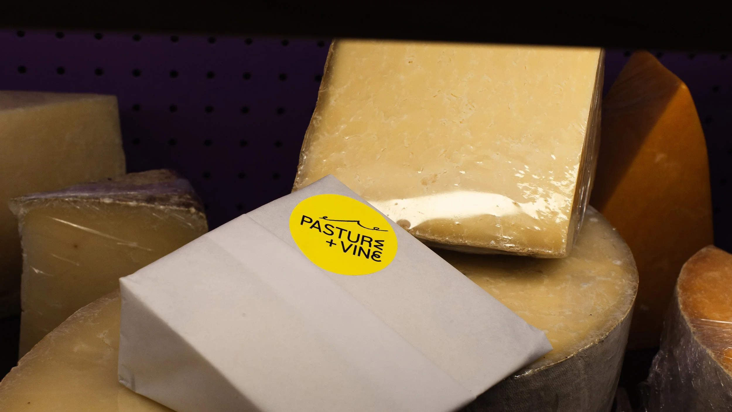 Round, yellow Pasture and Vine sticker on a piece of paper wrapped cheese sitting on a display of cut cheeses.
