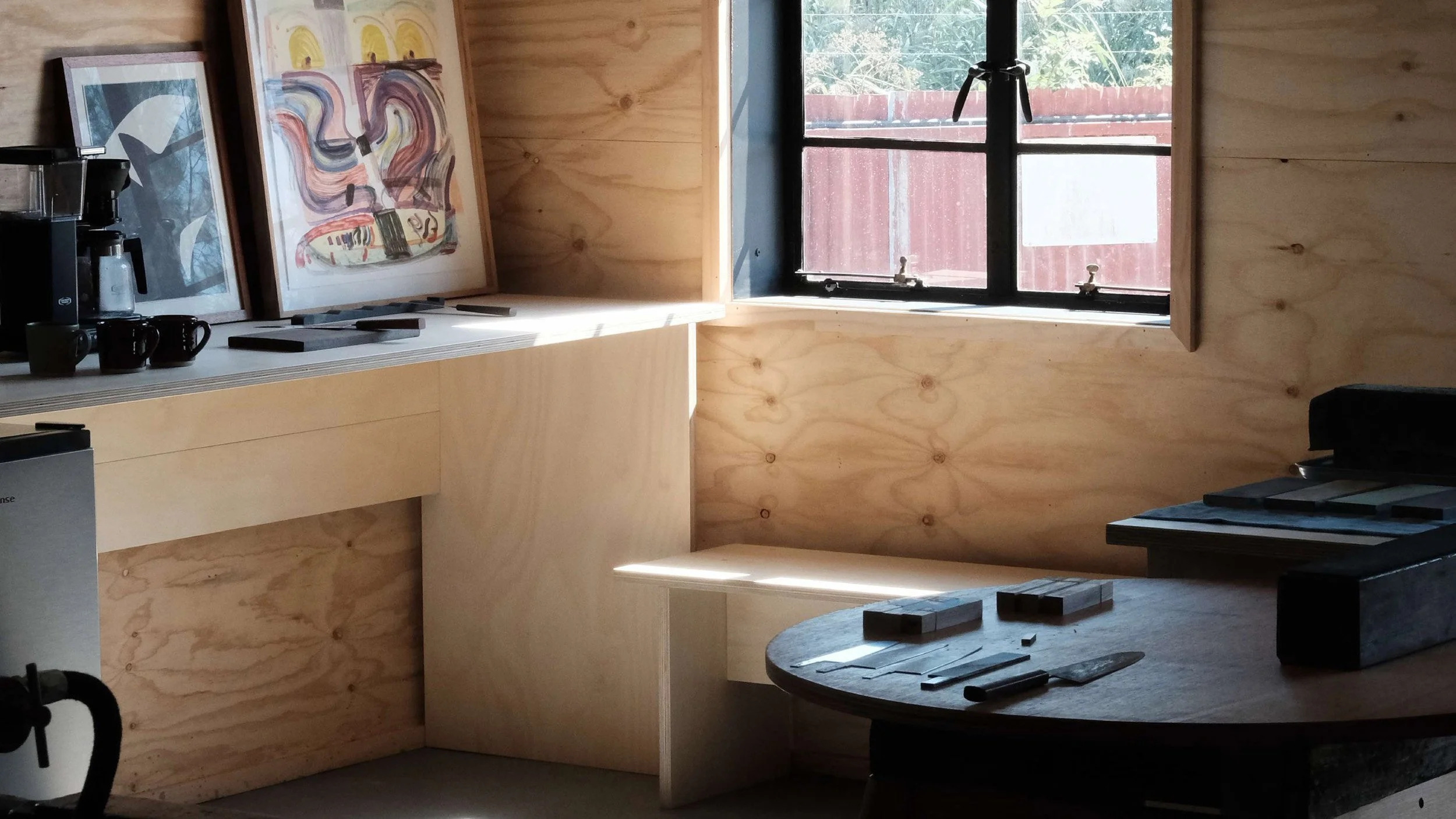Two Hands Forge workshop showing plywood clad walls and furniture. Knife blades and a complete knife sit on the table in the foreground.