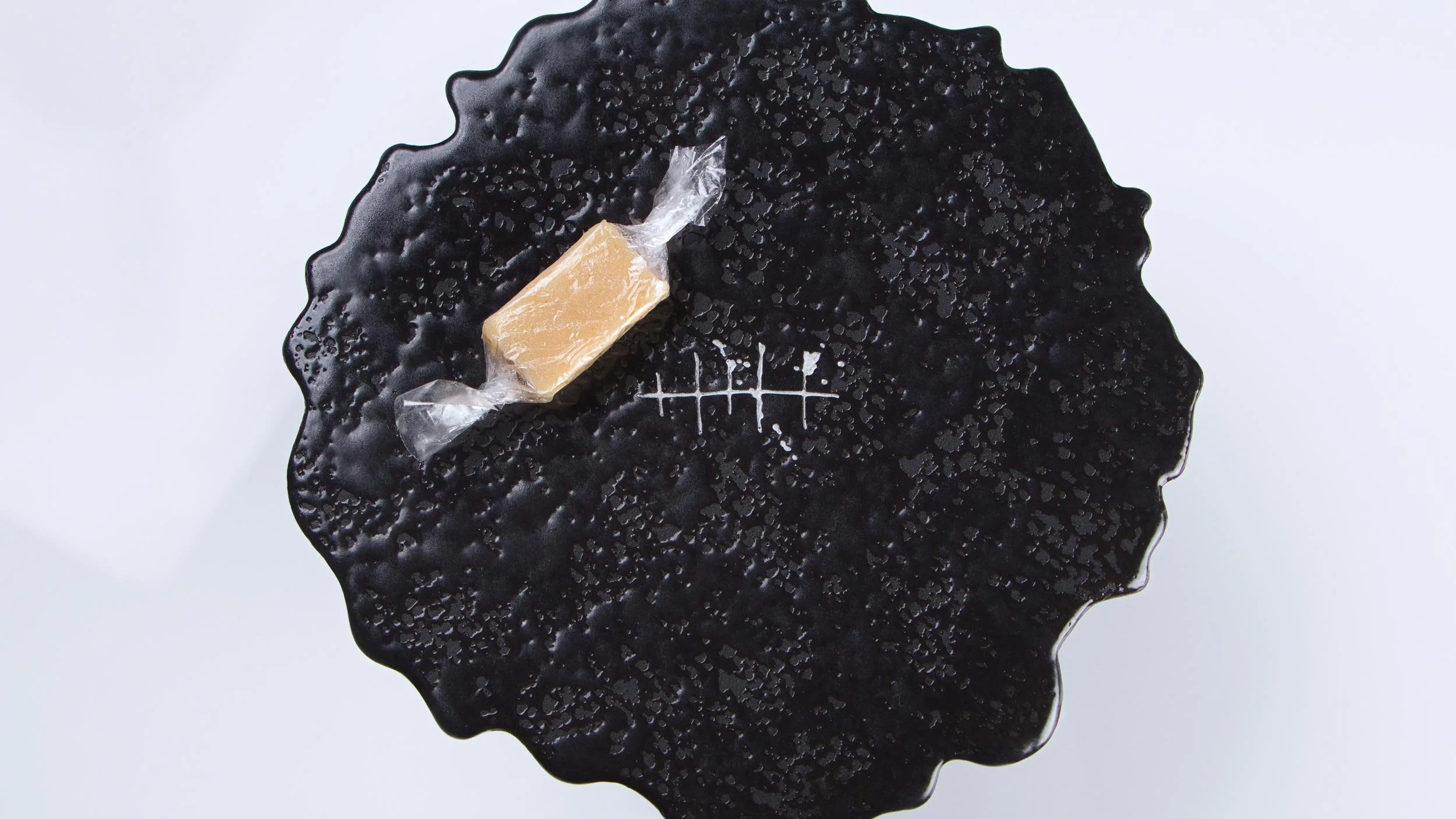 Black plate in the shape of an ink splat is engraved with the Tippling Club fishbone logo in white. One lolly in clear wrapping sits on top.