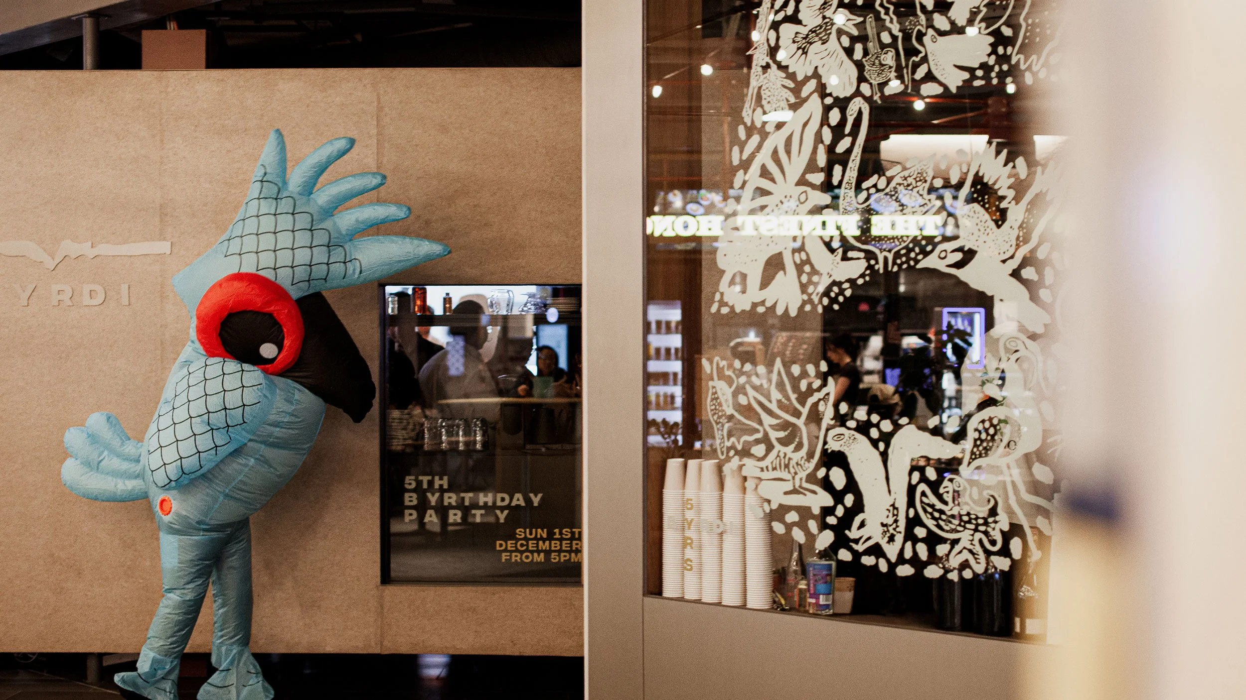 A person in an inflatable blue parrot costume stands in front of the entrance of Byrdi bar. In the foreground there is a decal on the window made up of illustrated birds in a 5 shape.