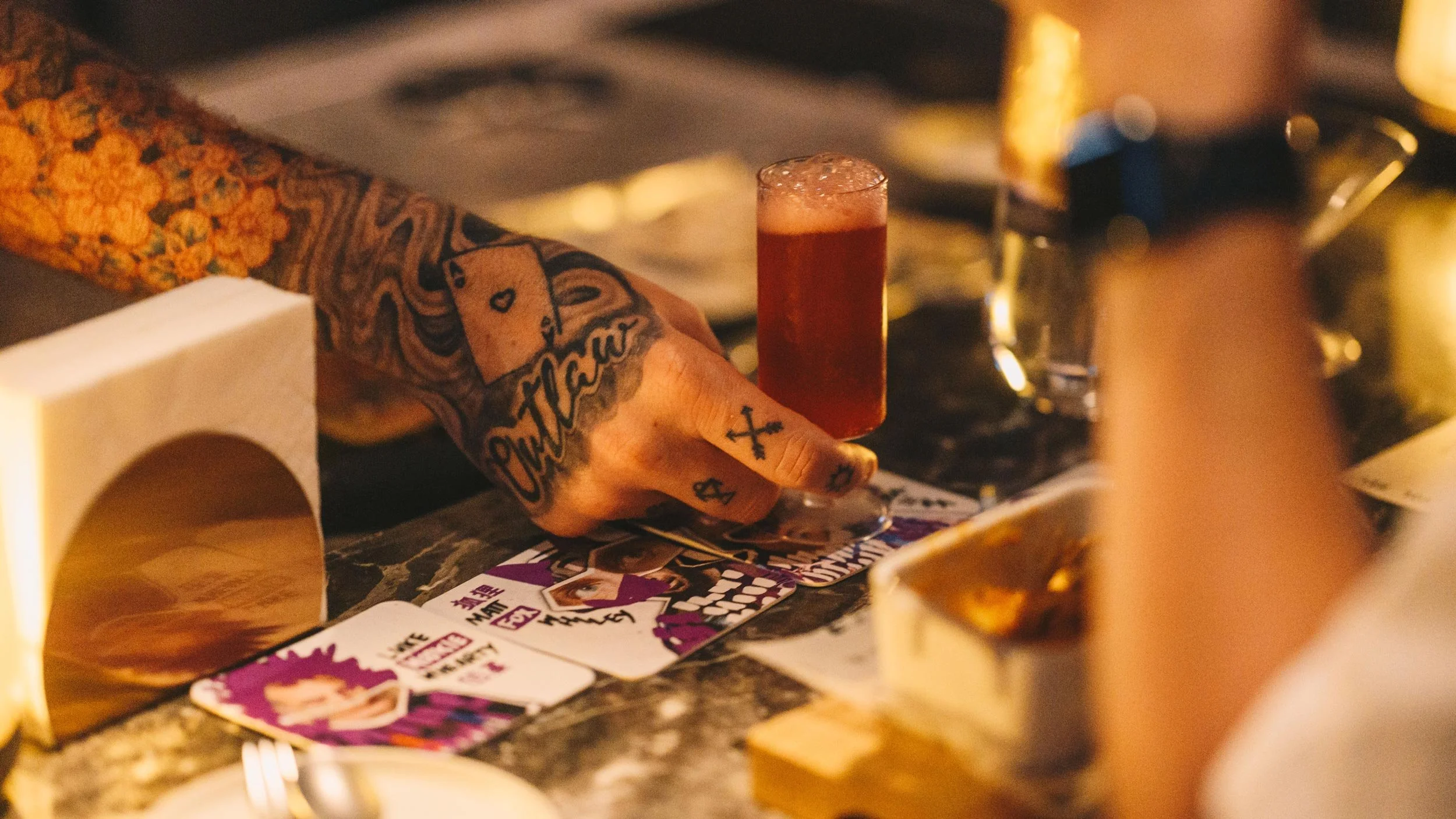 Tattooed hand of Matt Whiley serving up a cocktail on a LMC coaster. Two other coasters featuring LMC graphics sit alongside.
