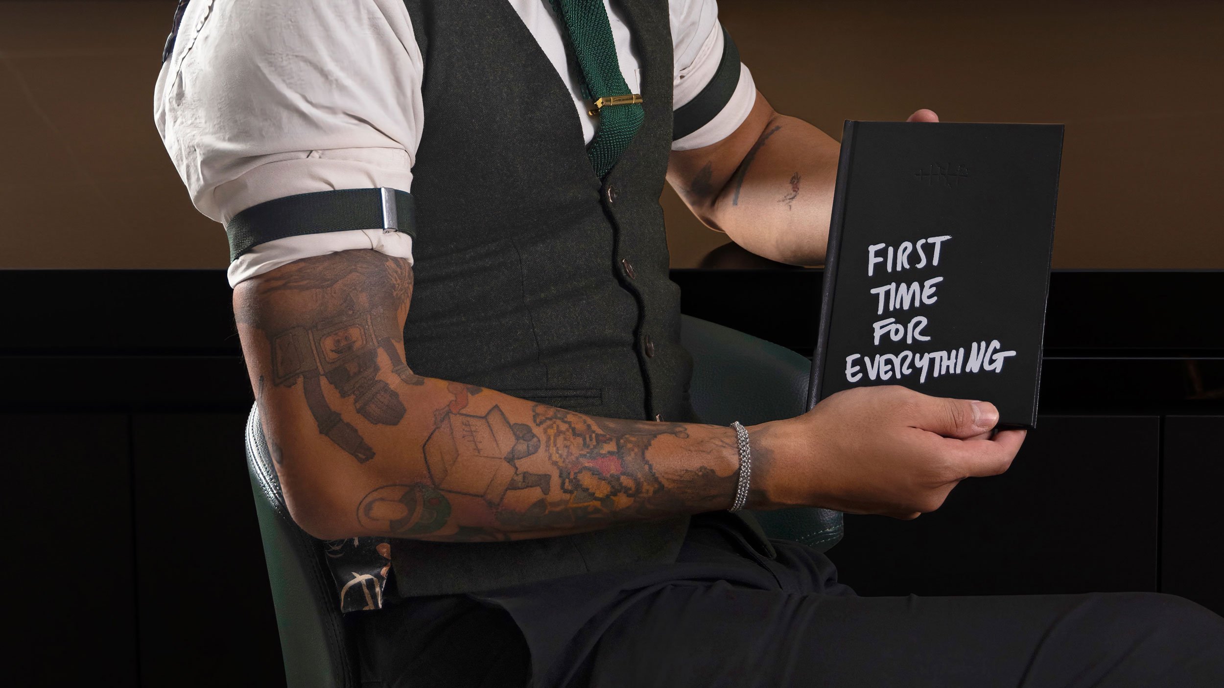 Arathorn Grey seated holding Tippling Club’s “First Time for Everything” cocktail menu. White hand drawn sharpie text on a black menu in the style of a journal.