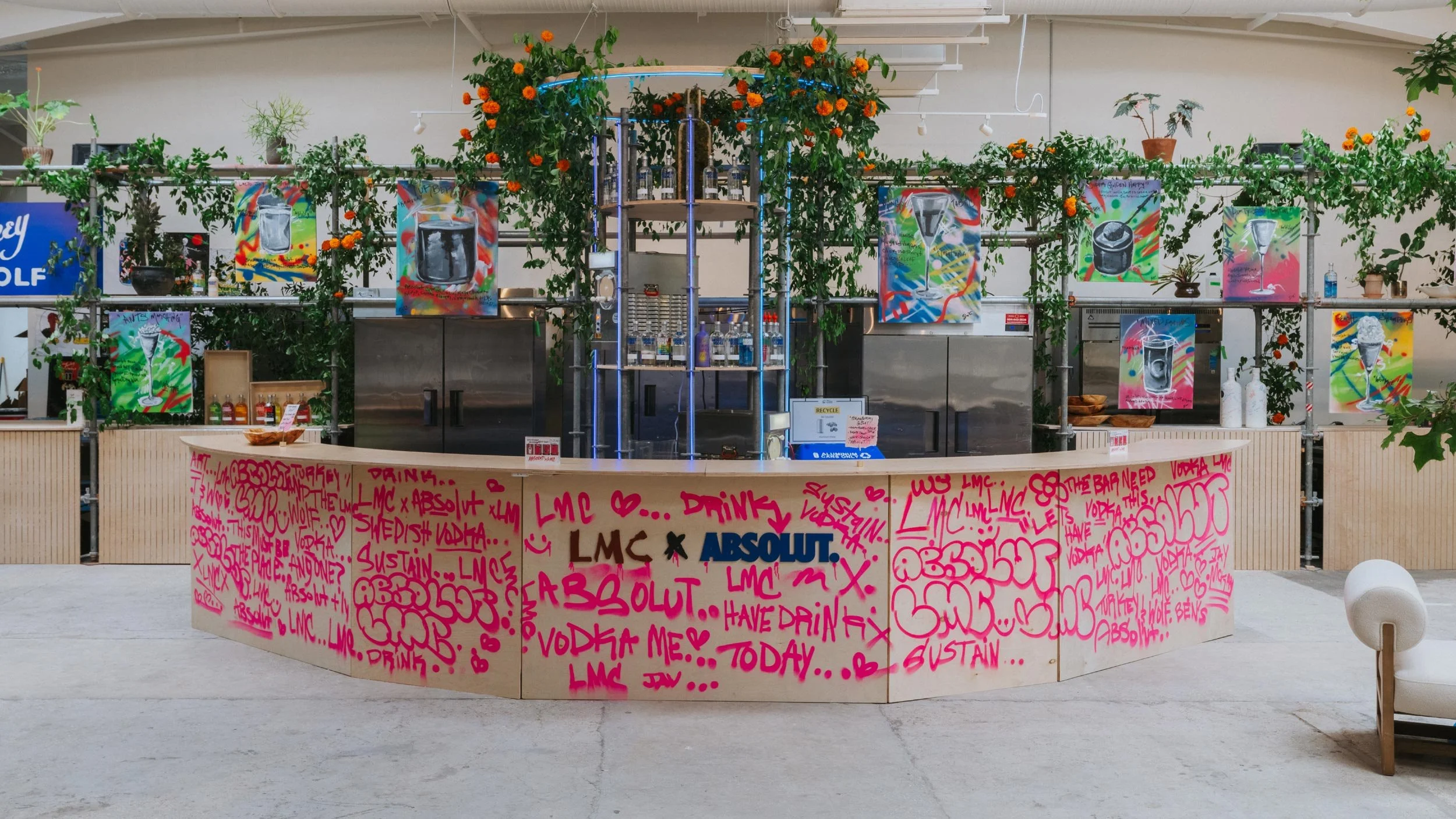 Wooden bar with LMC x Absolut logo lockup on the front. Pink spray painted words cover the bar. Behind the bar are large posters each with a cocktail spray painted on them forming a visual menu.