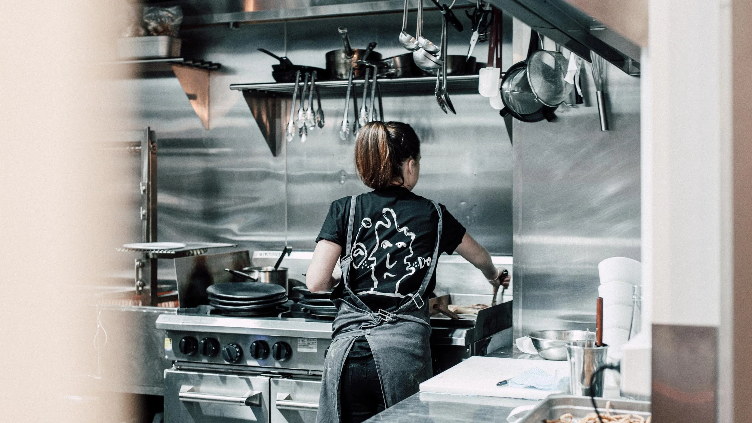 Rear view of chef in commercial kitchen tossing something over cooktop. Black Someday Sessions t-shirt with “three-headed drinker” illustration in white features.
