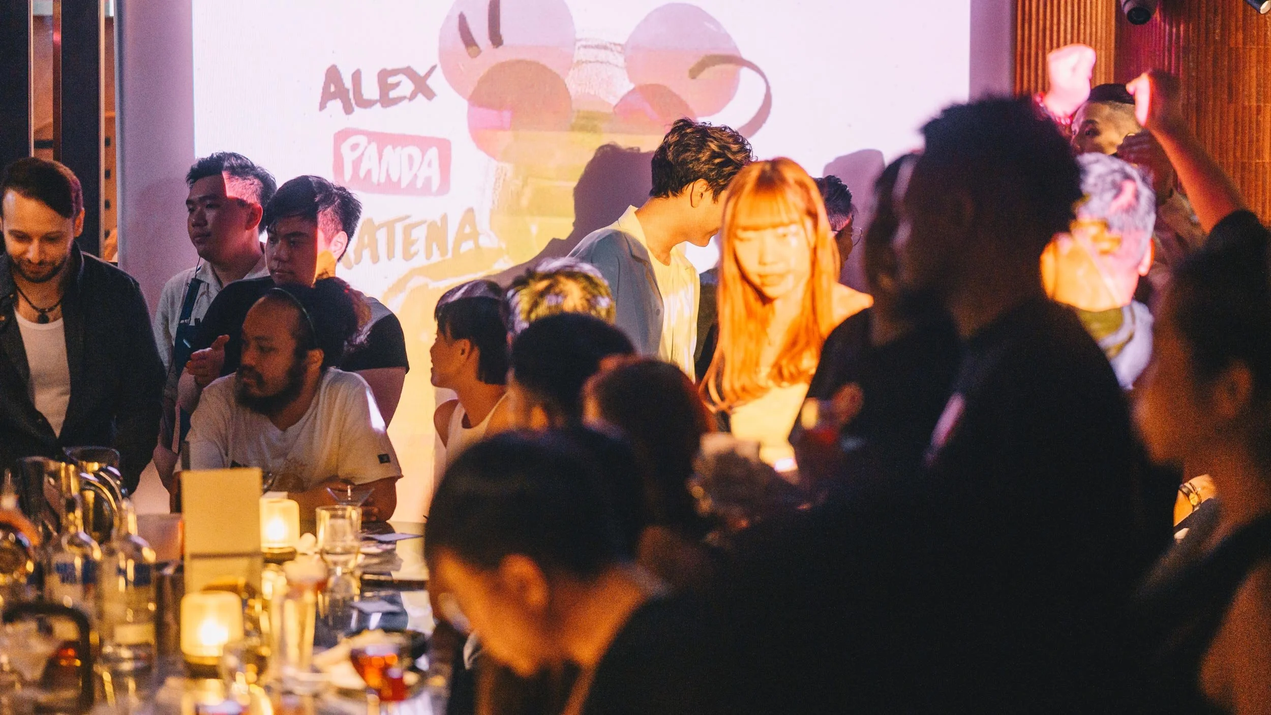 People mingling at The Opposites bar in Hong Kong during the LMC Vol #21 event. A projection screen is in the background showing a collage and type of Alex Panda Kratena.