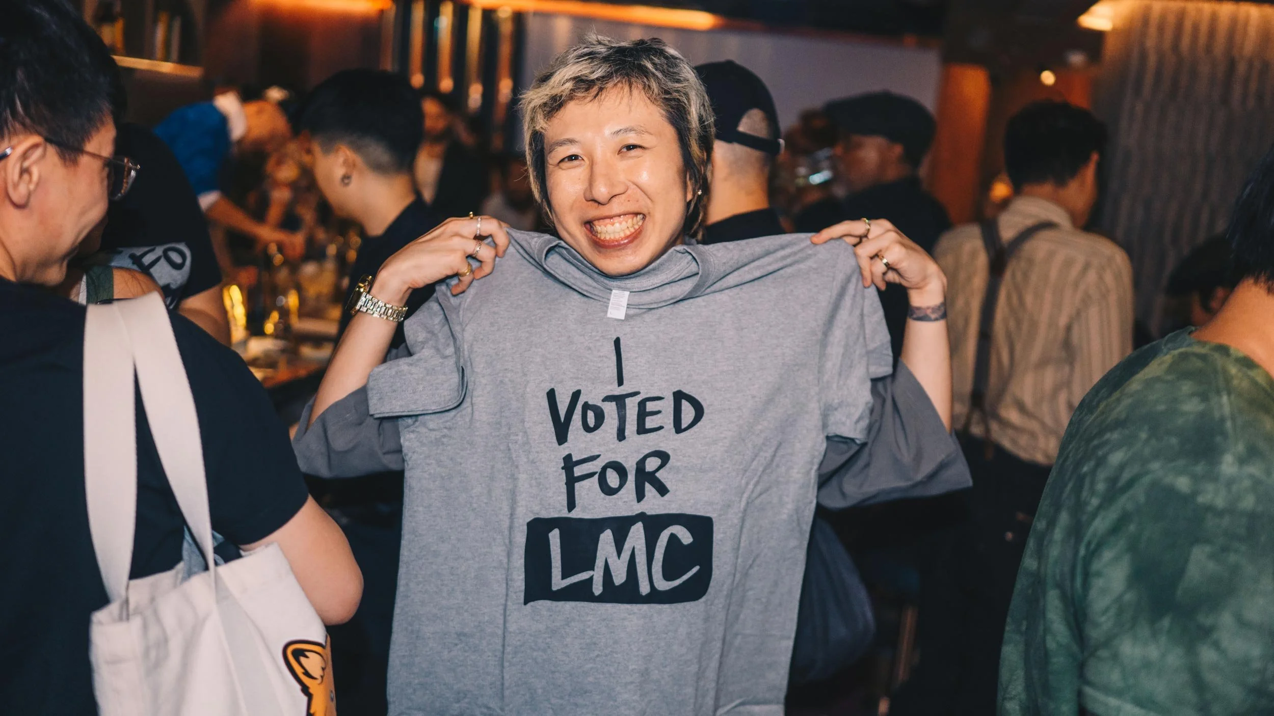 Short-haired smiling woman holding up a grey “I VOTED FOR LMC” t-shirt up against her torso posing to camera.