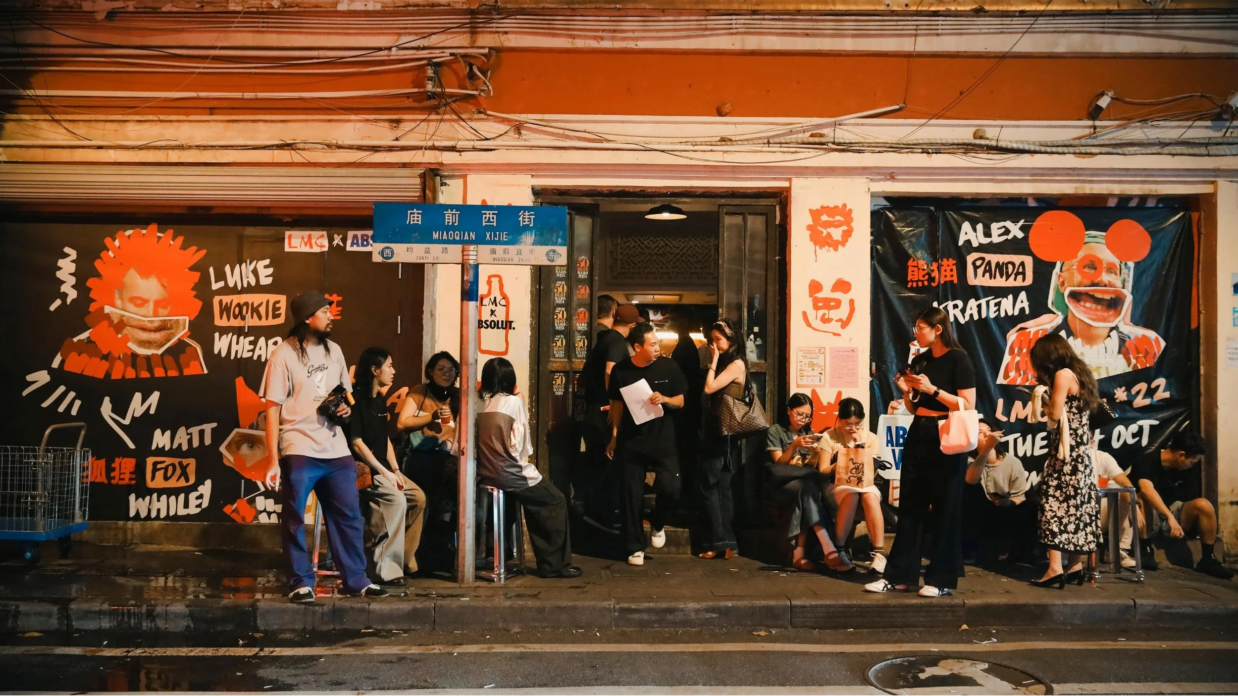 People mingling outside Hope & Sesame bar in Guangzhou, China. Graphics in black, white and orange promoting the LMC Vol #22 event cover the outside windows and pillars.