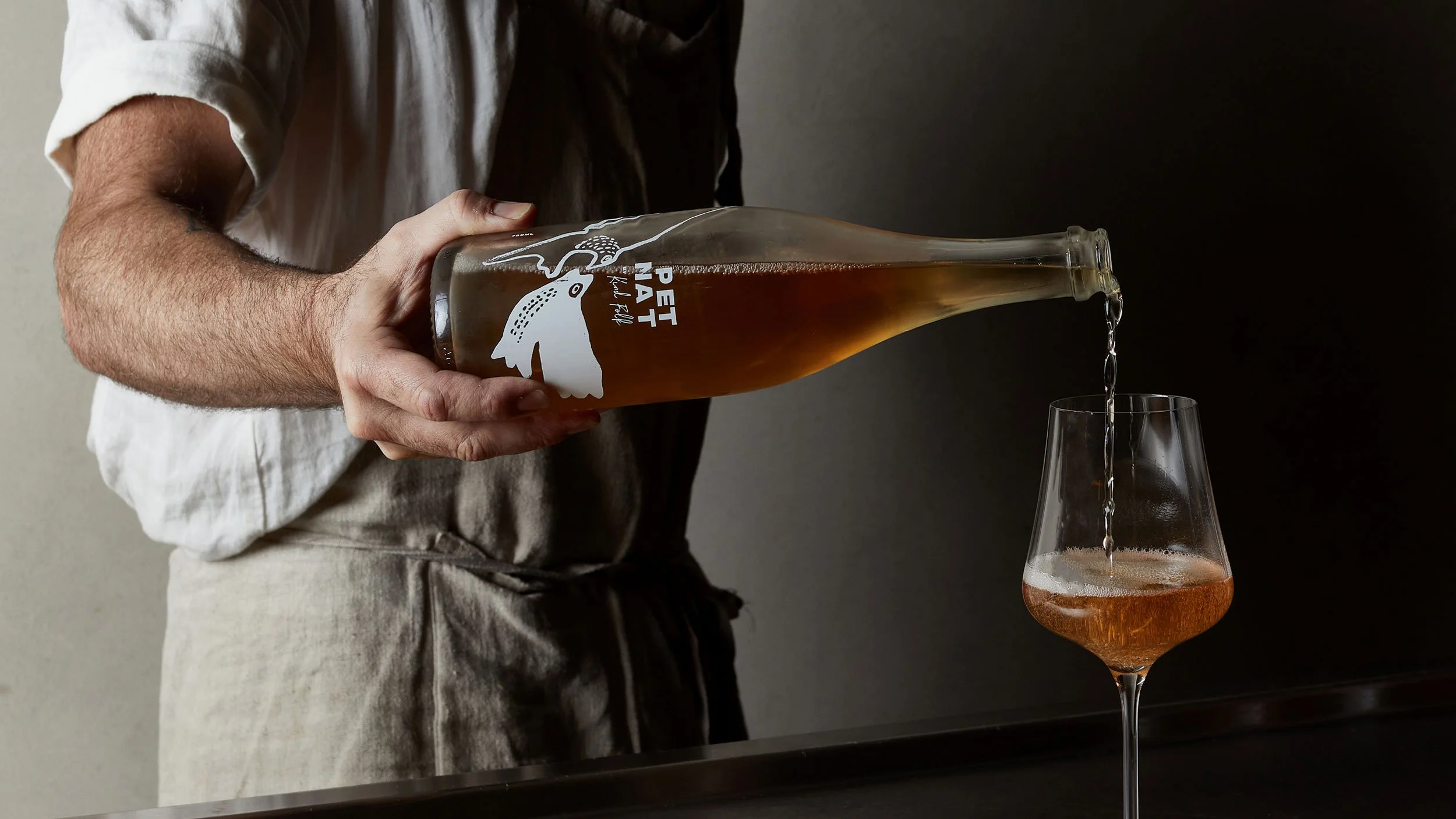 Person pouring a Byrdi x Kind Folk Pet Nat into a glass. On the bottle are illustrations of two welcome swallows.