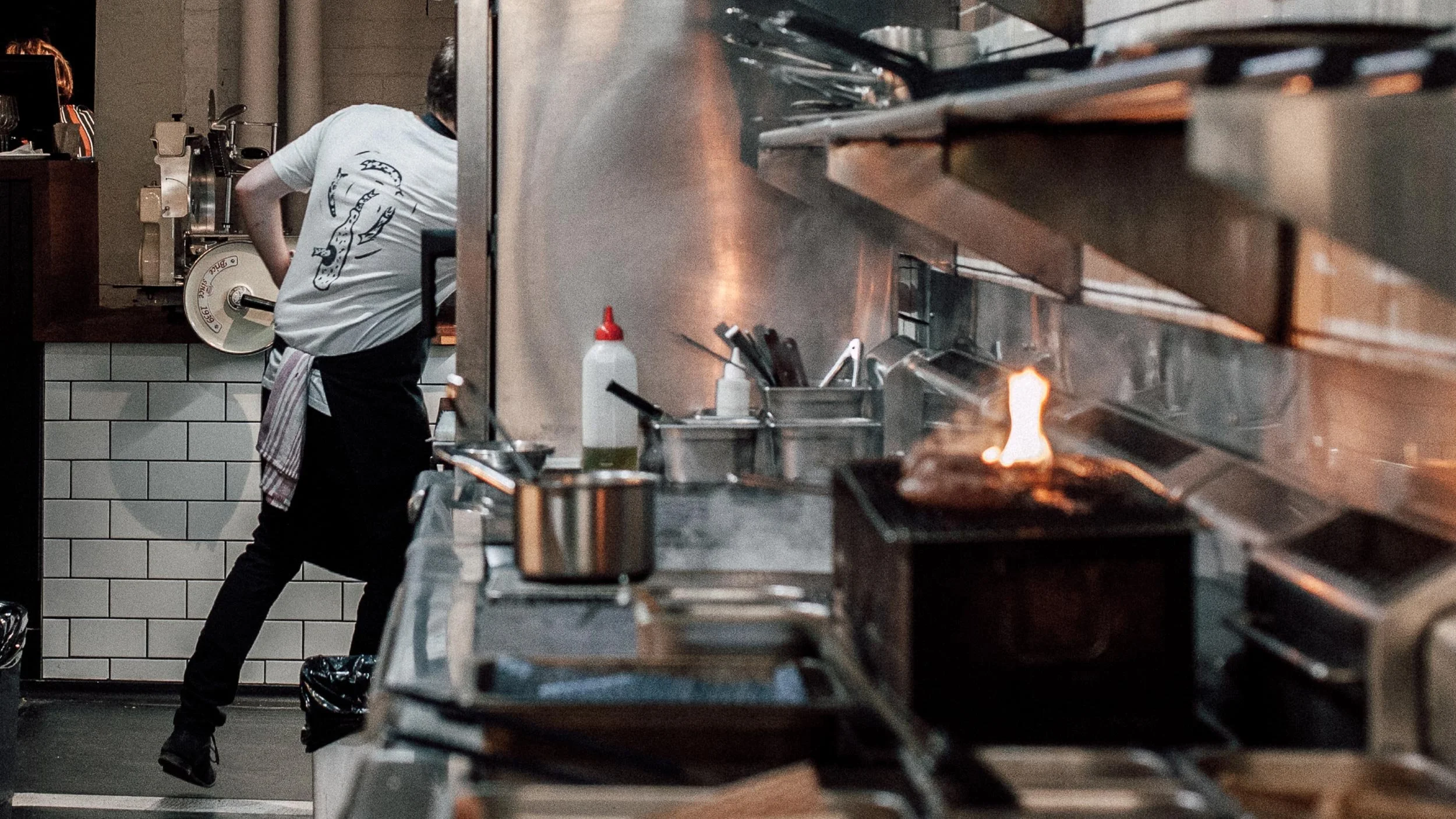 Rear view of Terry Dempsey running into commercial kitchen with meat on a griller in the foreground. White Someday Sessions t-shirt with with three anchovies circling around it in black features.