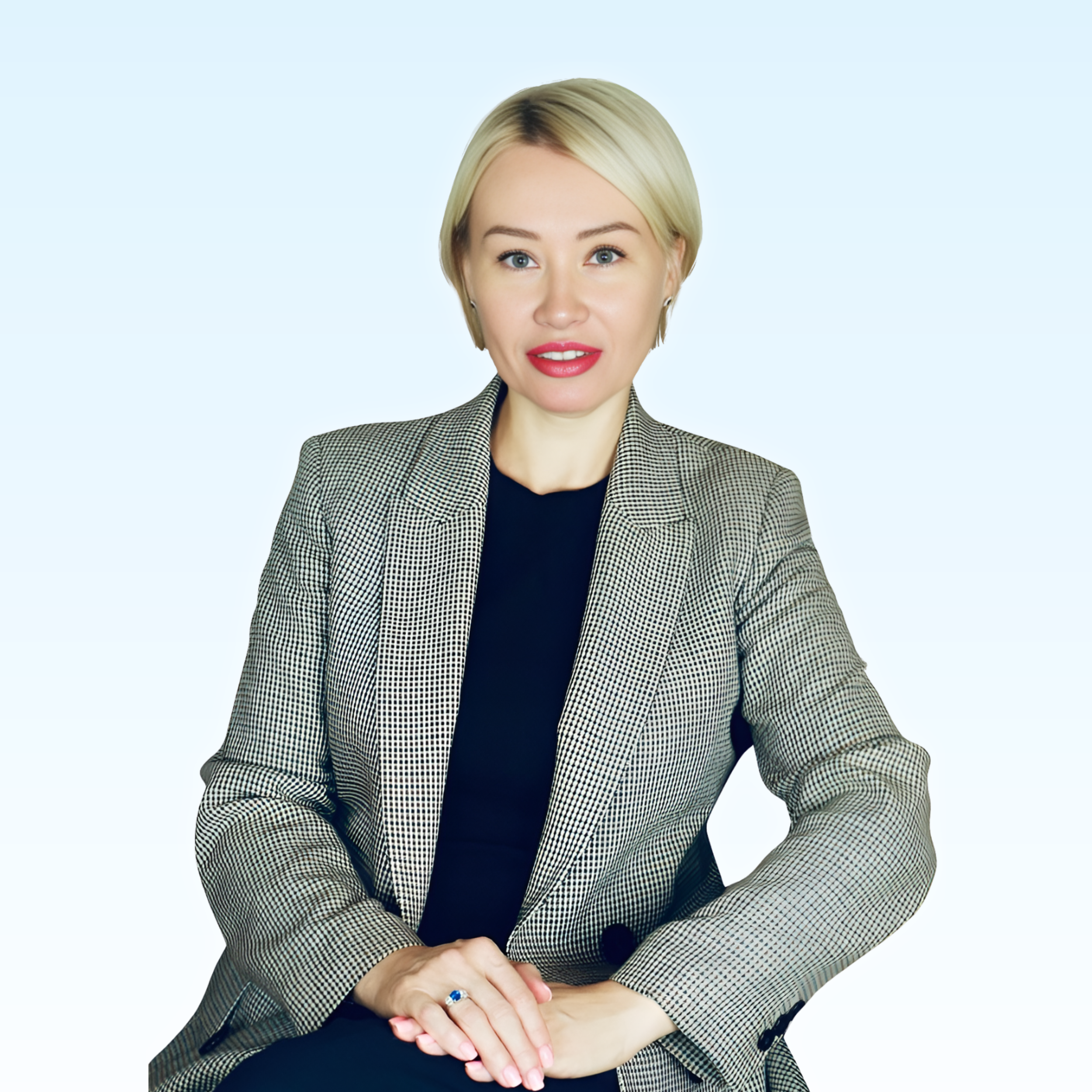 A woman with blonde hair, wearing a gray checked blazer and black top, sitting against a plain light background.