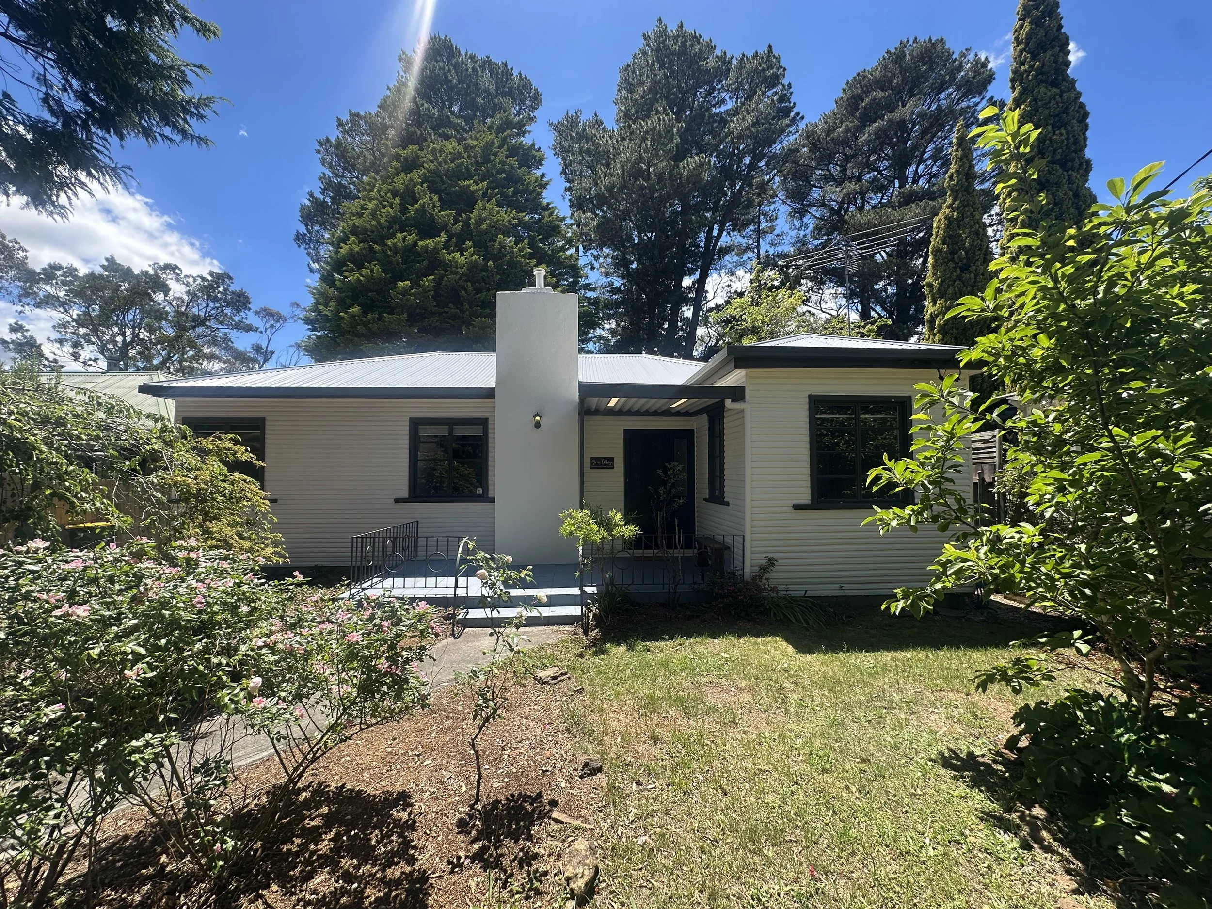 Exterior view of a white house with three windows, a door with a screen door, and an air conditioning unit outside. The house has a small porch with steps and a railing. The sky is partly cloudy with blue sky and fluffy clouds.