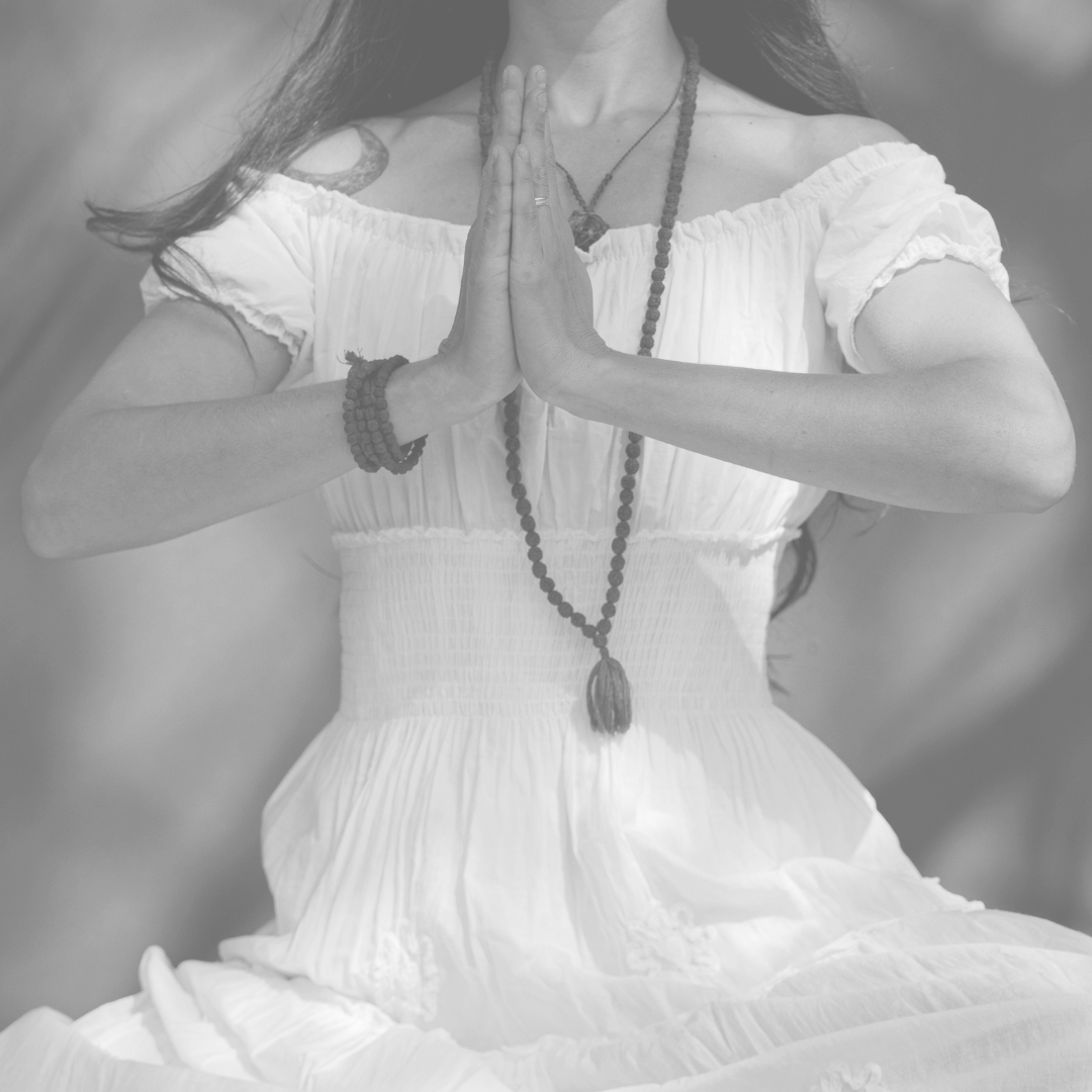 une femme en robe blanche pratique la méditation, mains jointes en symbole de recueillement, portant plusieurs bracelets et un collier avec une amulette