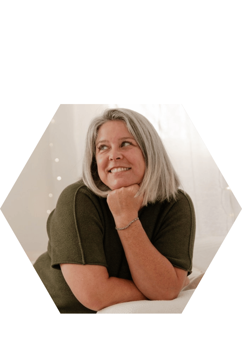 Une femme aux cheveux gris, souriante, repose sa tête sur sa main, dans une pièce lumineuse.