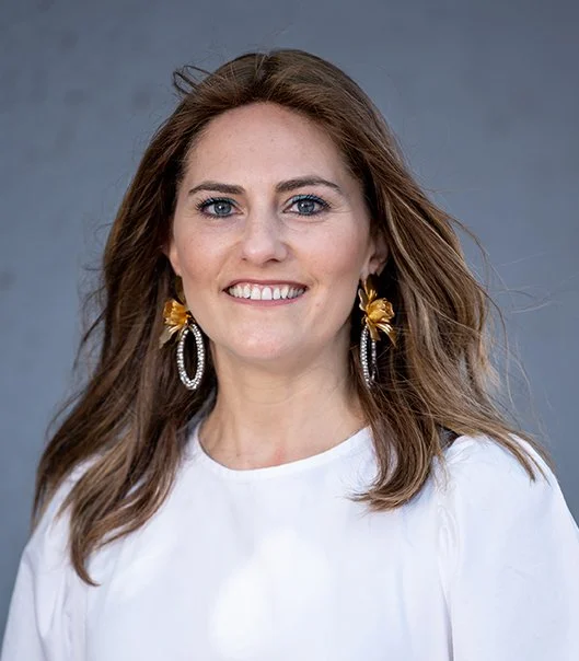A woman with long brown hair wearing a white top and large gold and silver earrings, smiling at the camera against a gray background.