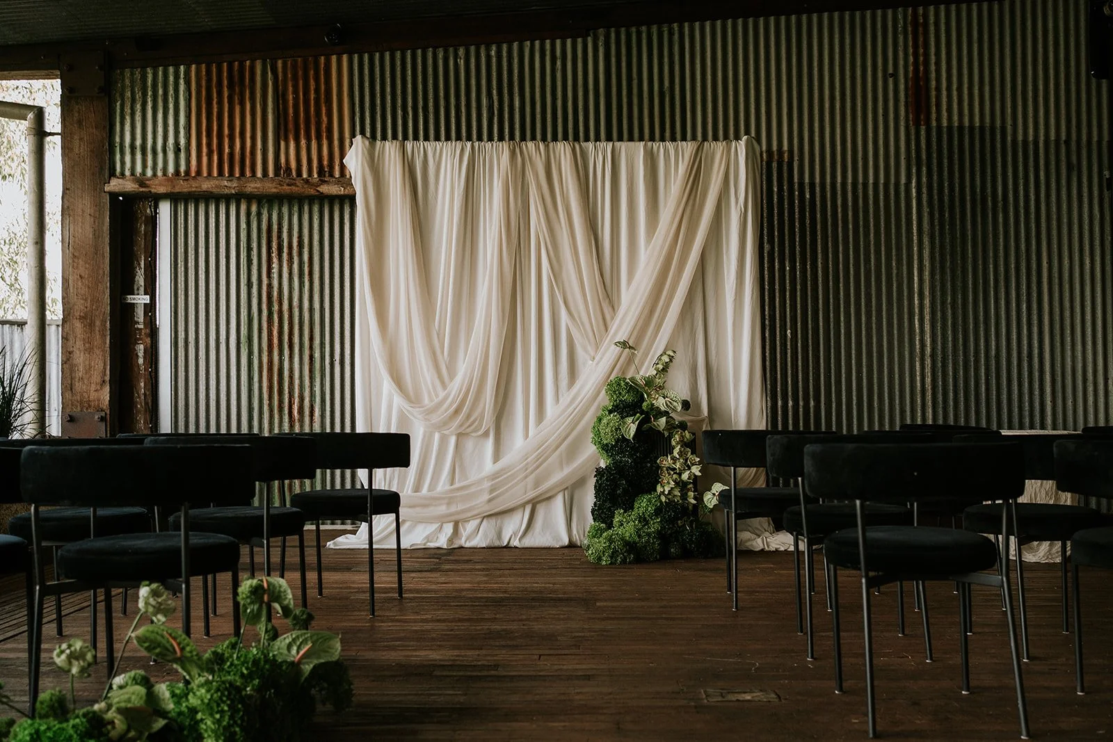 Indoor wedding ceremony setup with black chairs, white draped fabric backdrop, greenery arrangement, and a rustic corrugated metal wall.