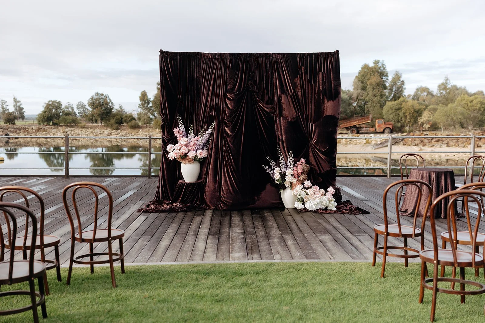 Outdoor wedding ceremony setup with a dark velvet fabric backdrop decorated with pink and white floral arrangements, wooden chairs, and a scenic water view in the background.