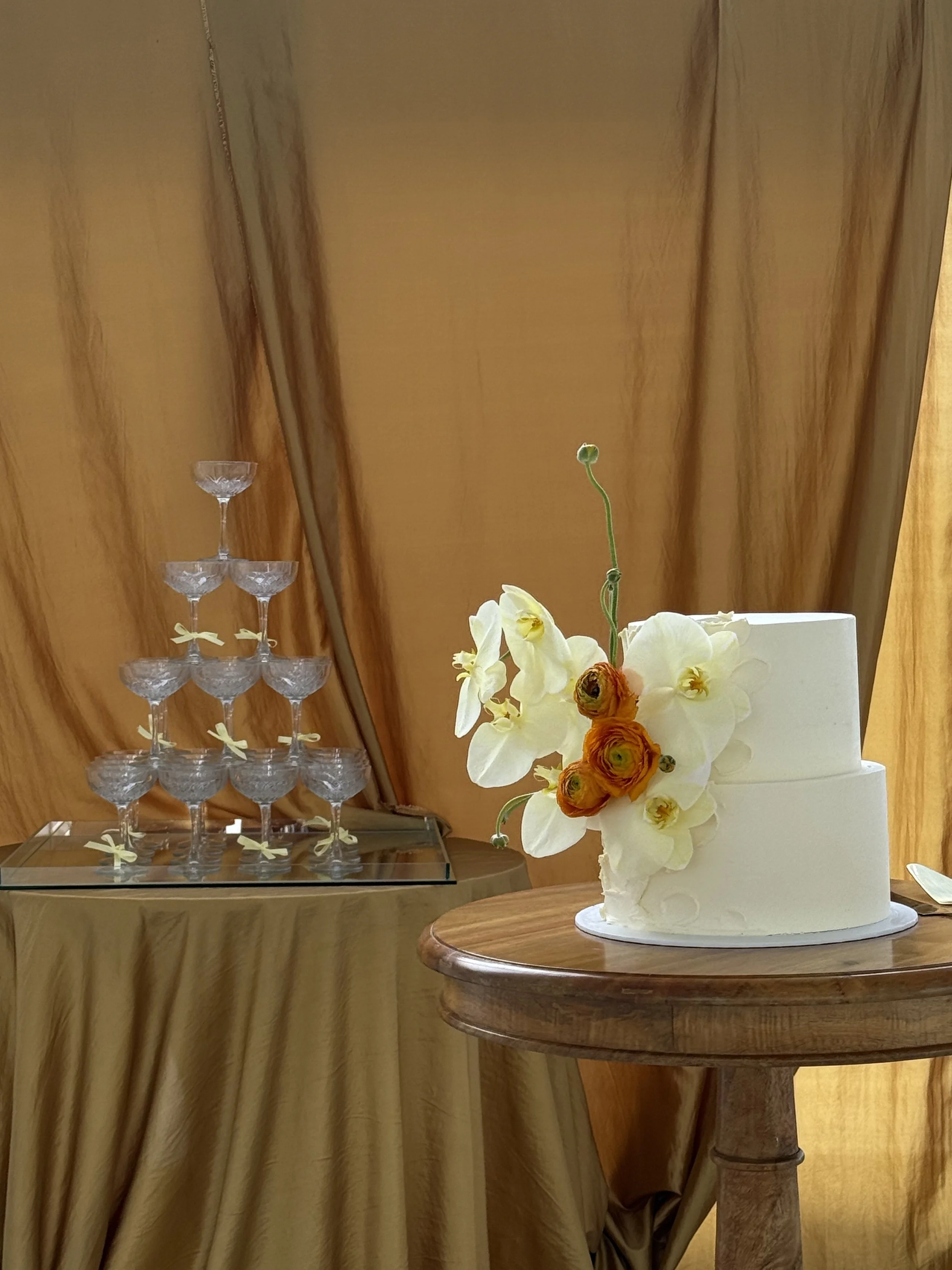 White wedding cake decorated with white and orange flowers on a wooden table with a champagne tower in the background.