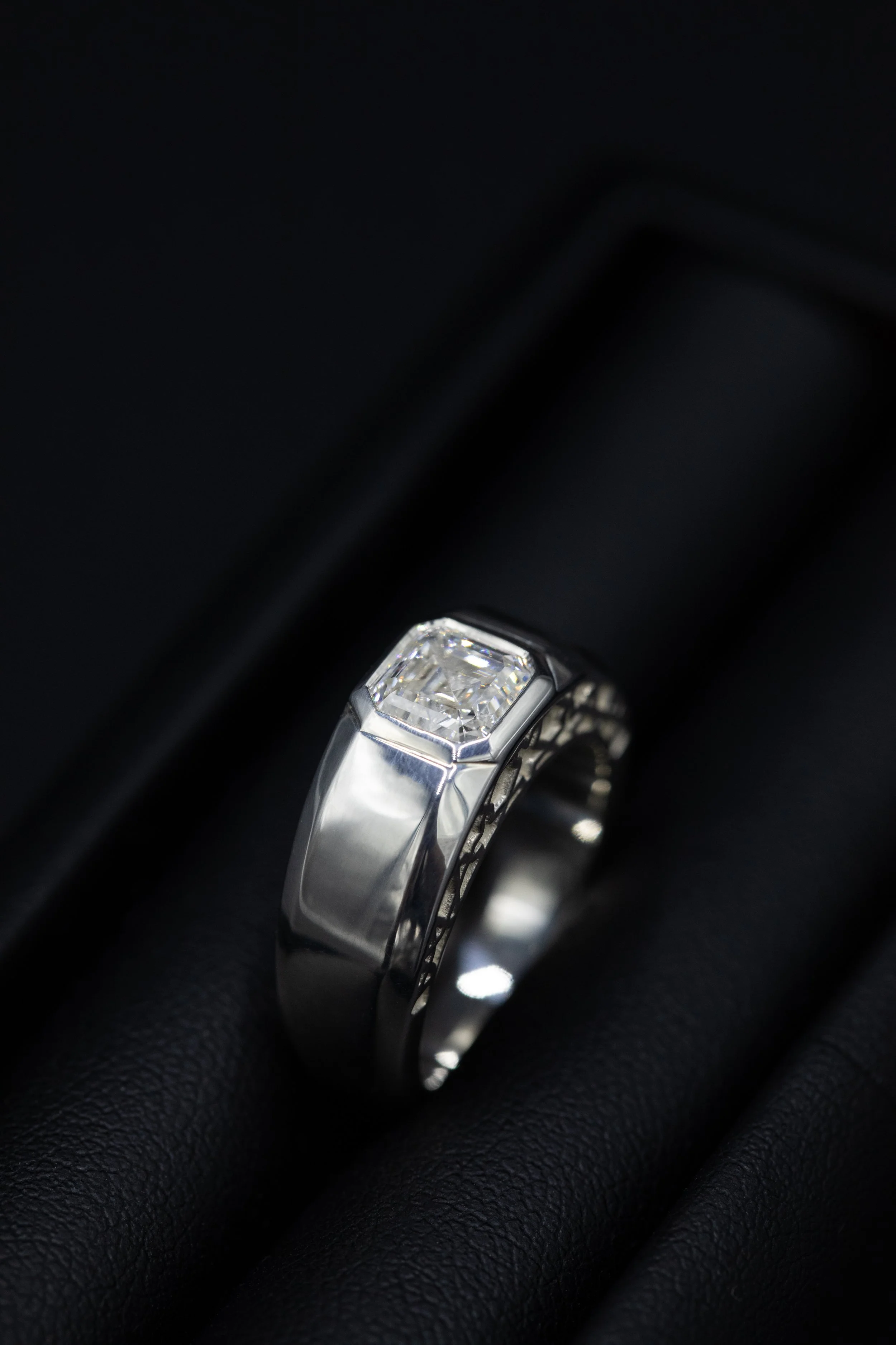 A silver men's ring with a square asscher cut diamond in the center, a geometric design on both sides resting on a black textured surface.