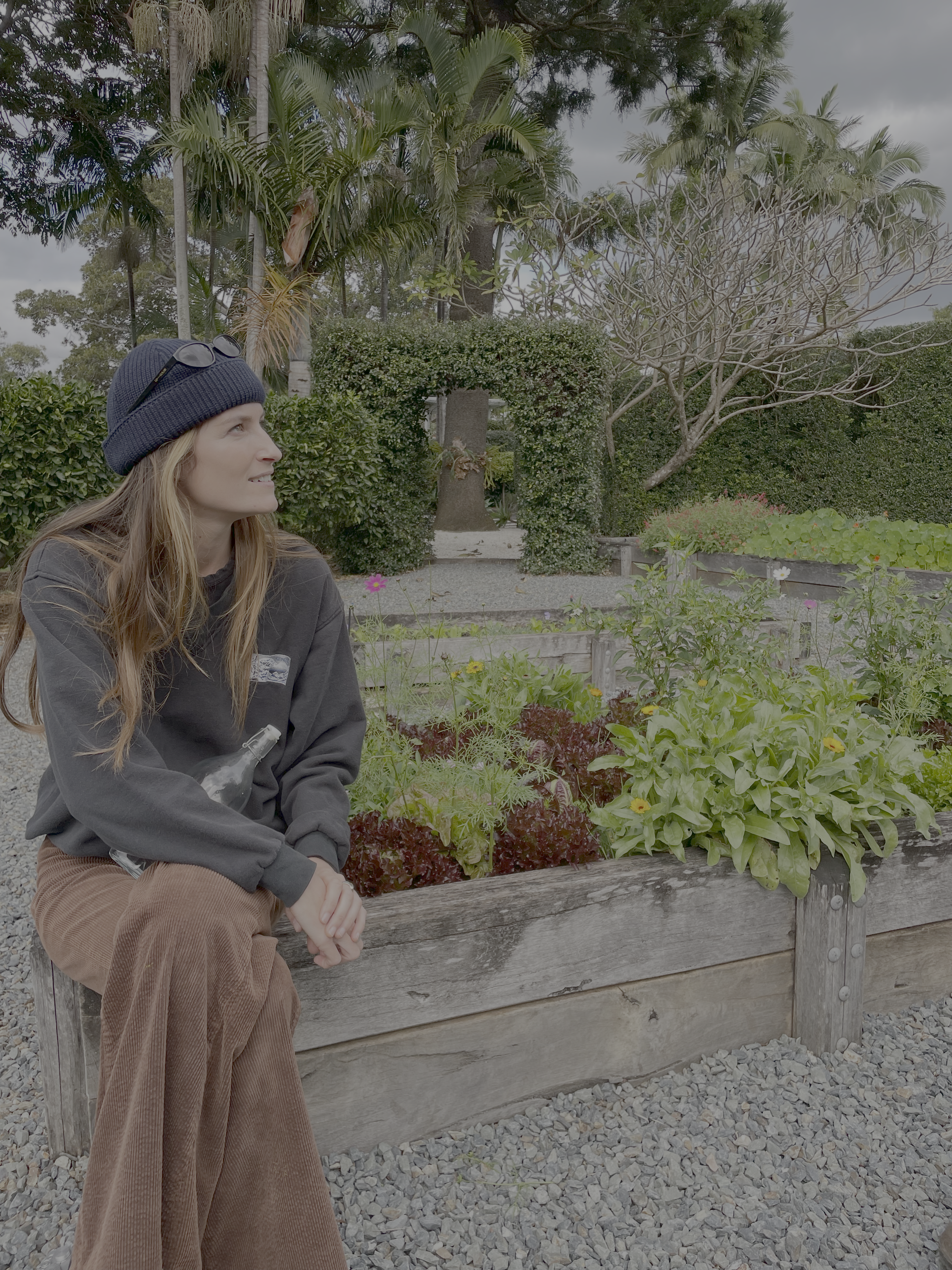 A young woman with long blonde hair, wearing a navy beanie with glasses on top, brown corduroy pants, and a dark jacket, sitting on a wooden garden bed filled with leafy green vegetables and flowers, looking to her left in an outdoor garden with tree
