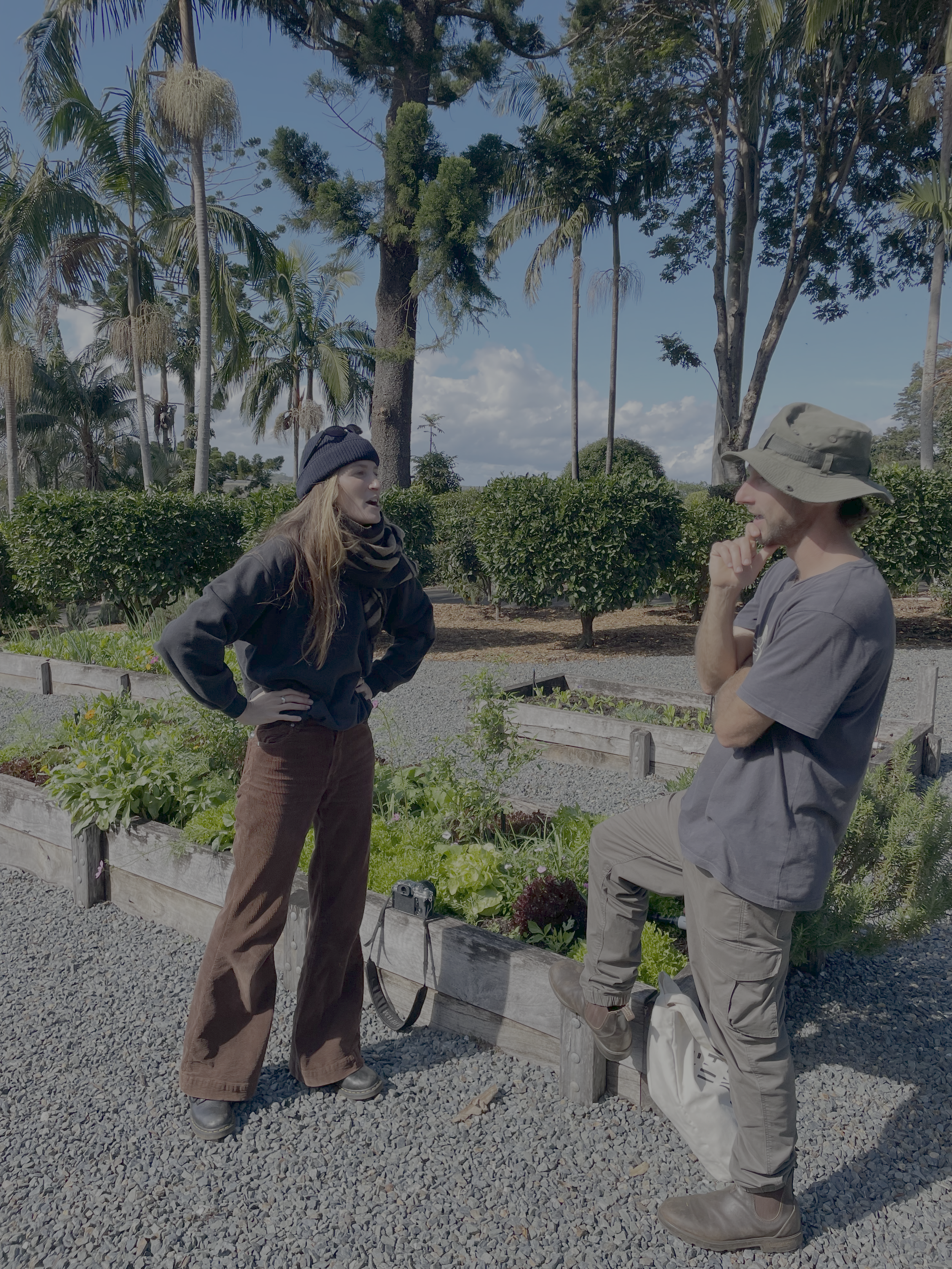 Two people, a woman and a man, are standing and talking in a garden with trees and plants. The woman is wearing a black beanie, black jacket, and brown pants, while the man is wearing a wide-brimmed hat, gray T-shirt, and tan pants. One of them has t