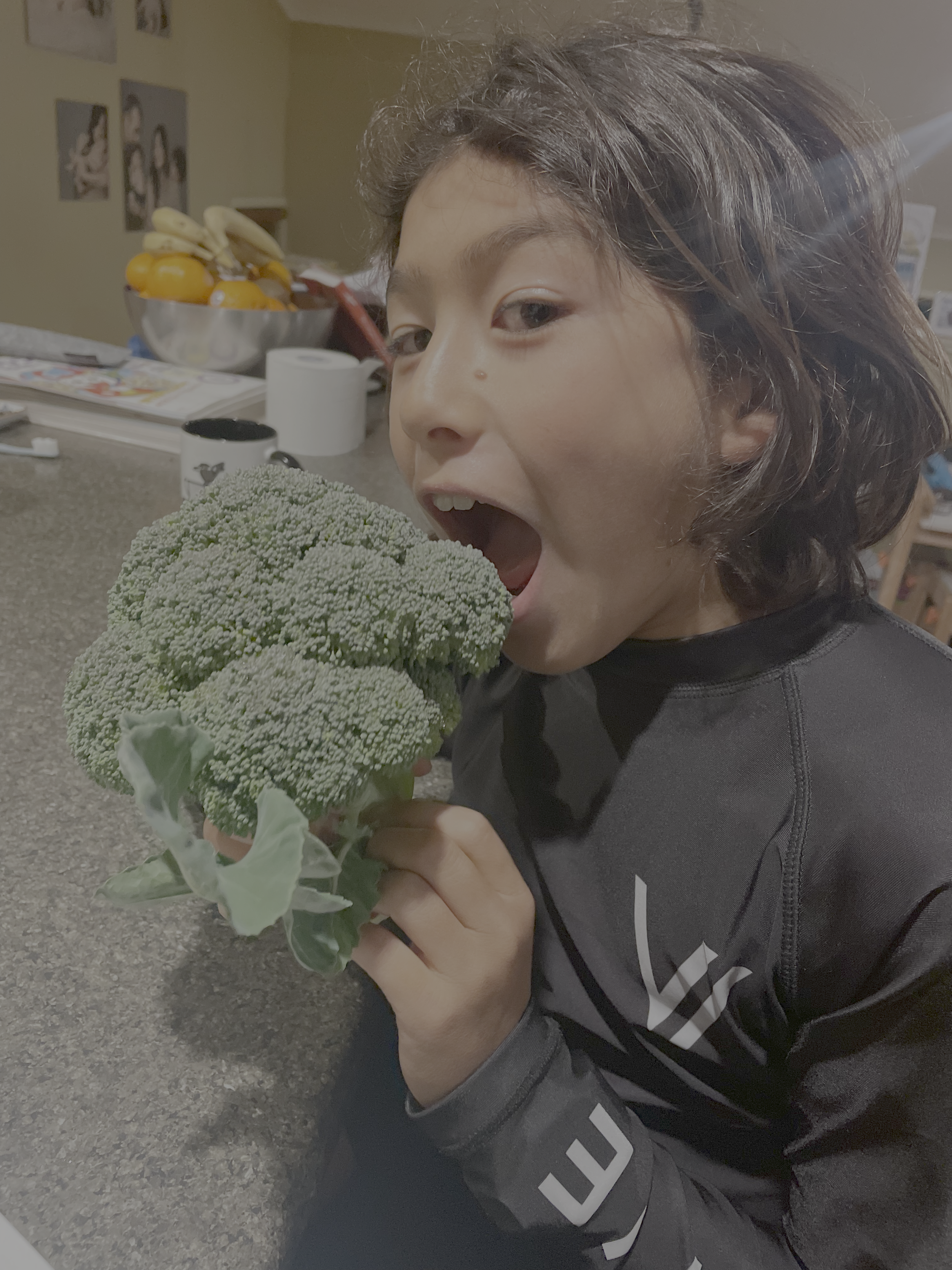 A young person with short, wavy dark hair and light skin is posing with an open mouth near a large green broccoli head in an indoor setting.