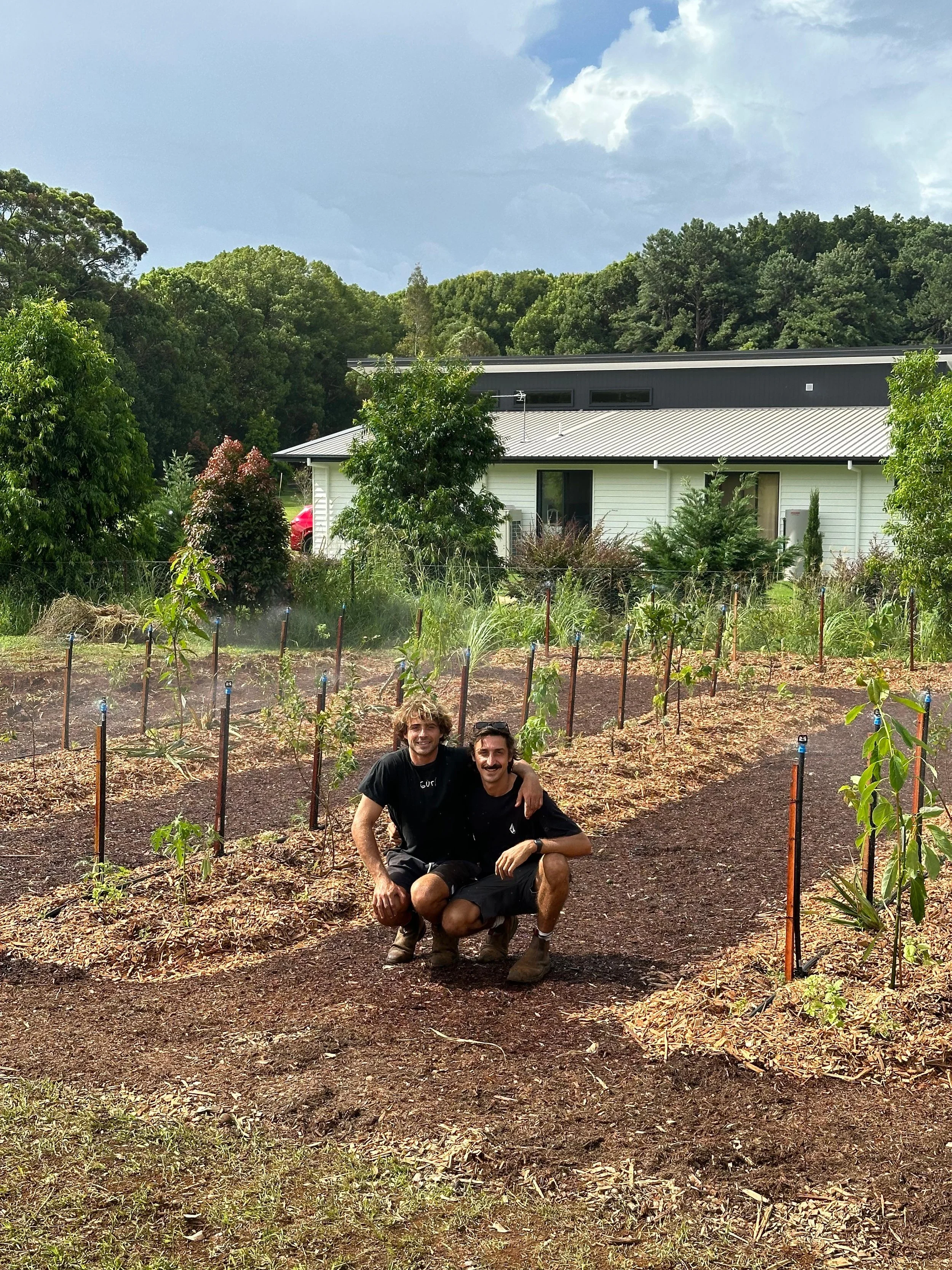 Two young men smiling and crouching in a garden or farm with young plants, surrounded by trees and a house in the background.