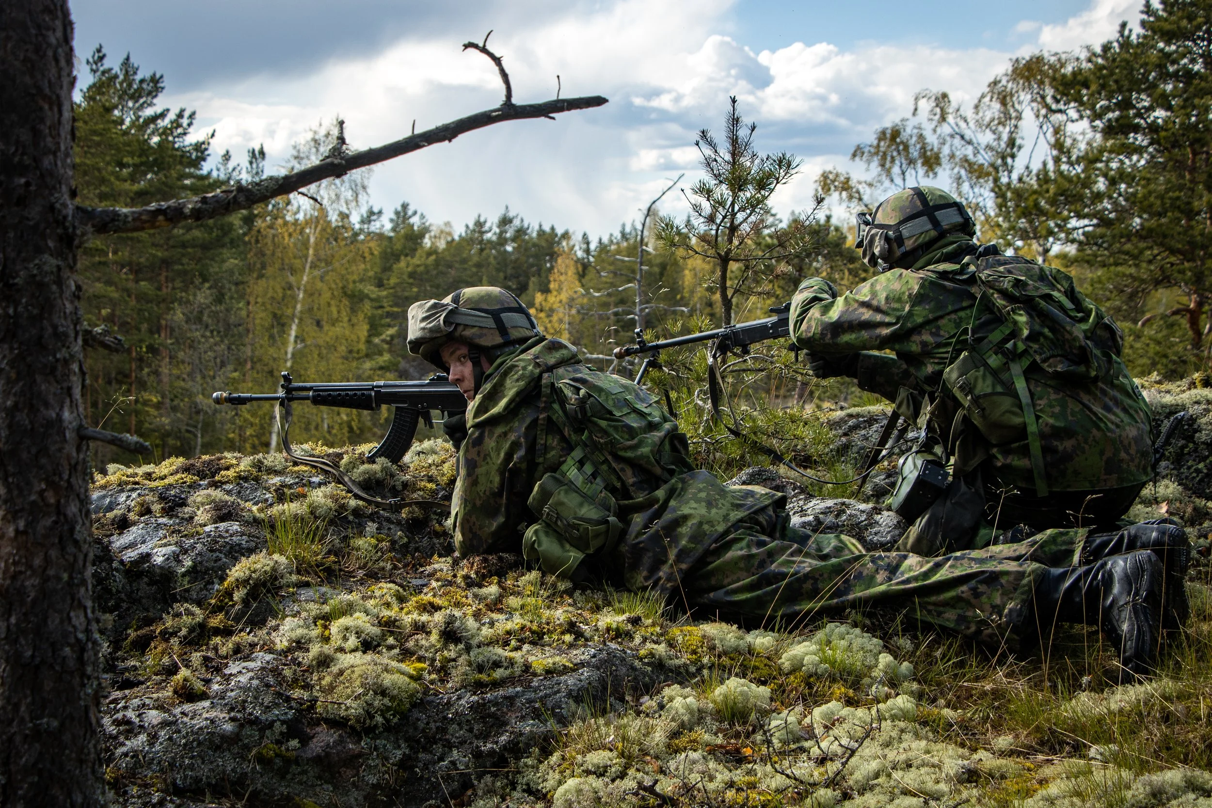 Two soldiers in camouflage gear lying prone in a forest, aiming rifles through the trees.
