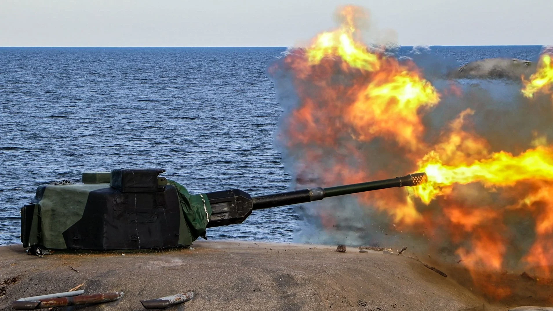 A military cannon firing a projectile with a large explosion and fire bursting out of the barrel, near the ocean.