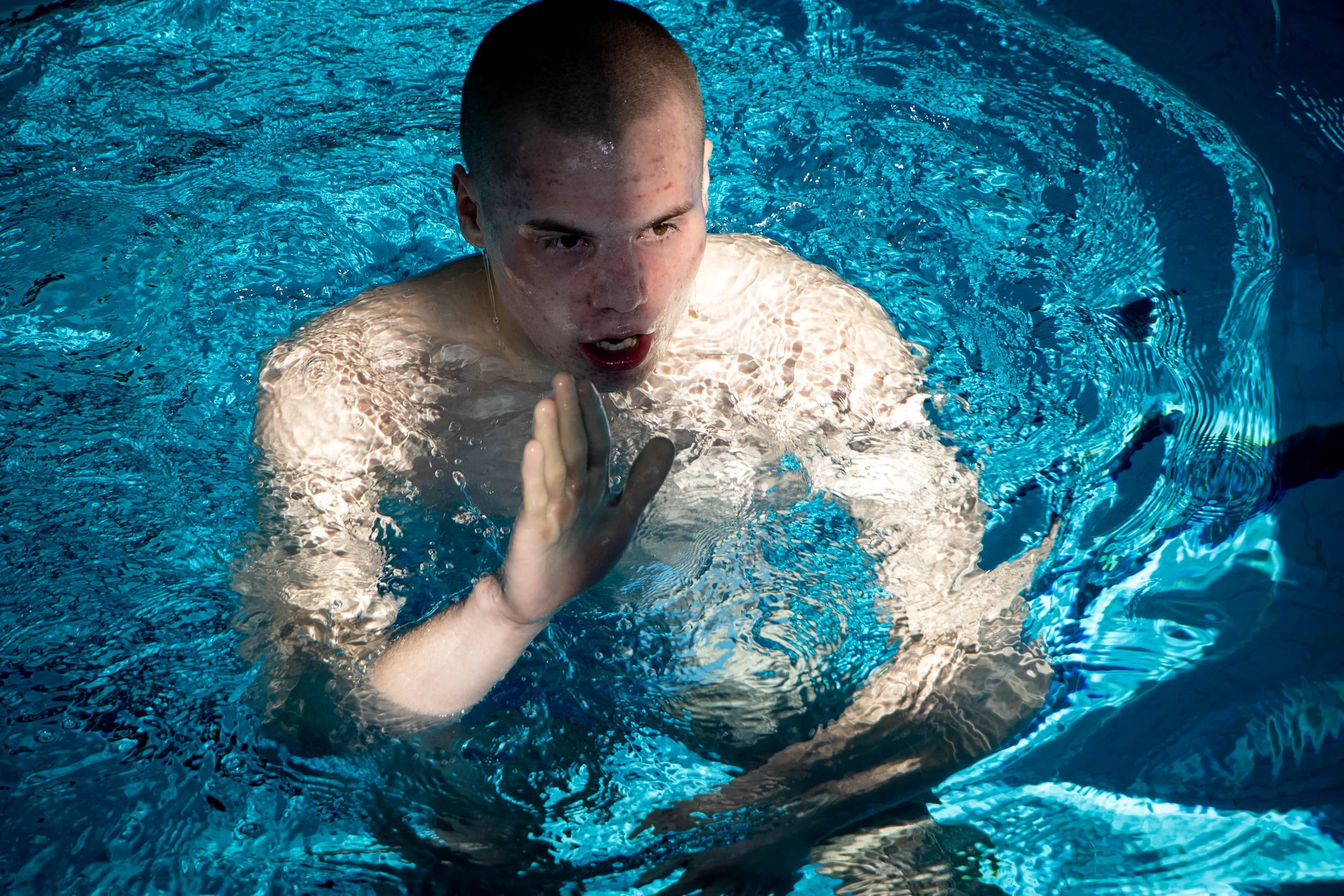 A young man with short hair is in a swimming pool, with water up to his chest. He has an intense expression on his face and is holding his right hand up, as if signaling or stopping. The water is clear blue, and there are ripples around him.