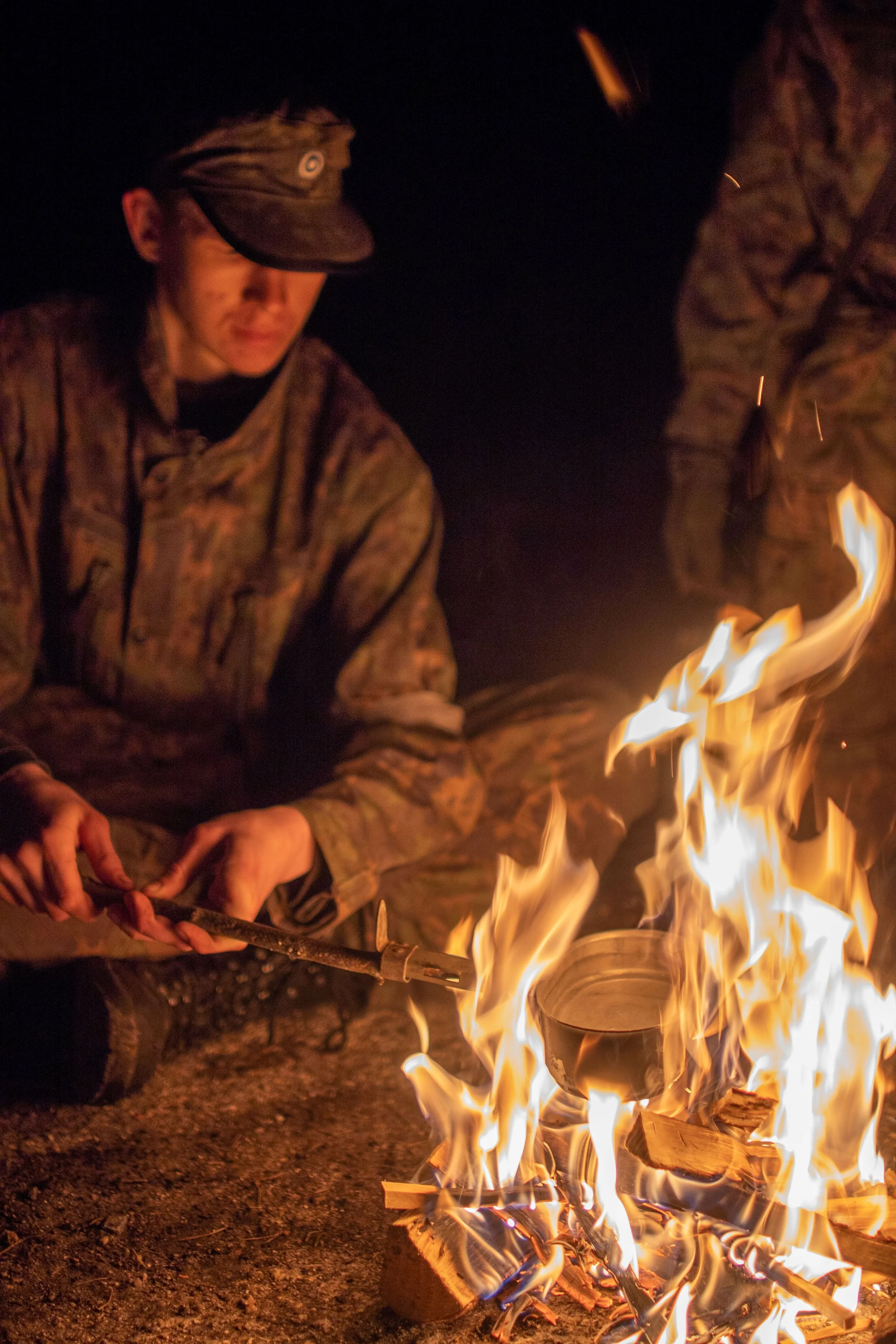 Person wearing camouflage clothing tending a campfire at night.