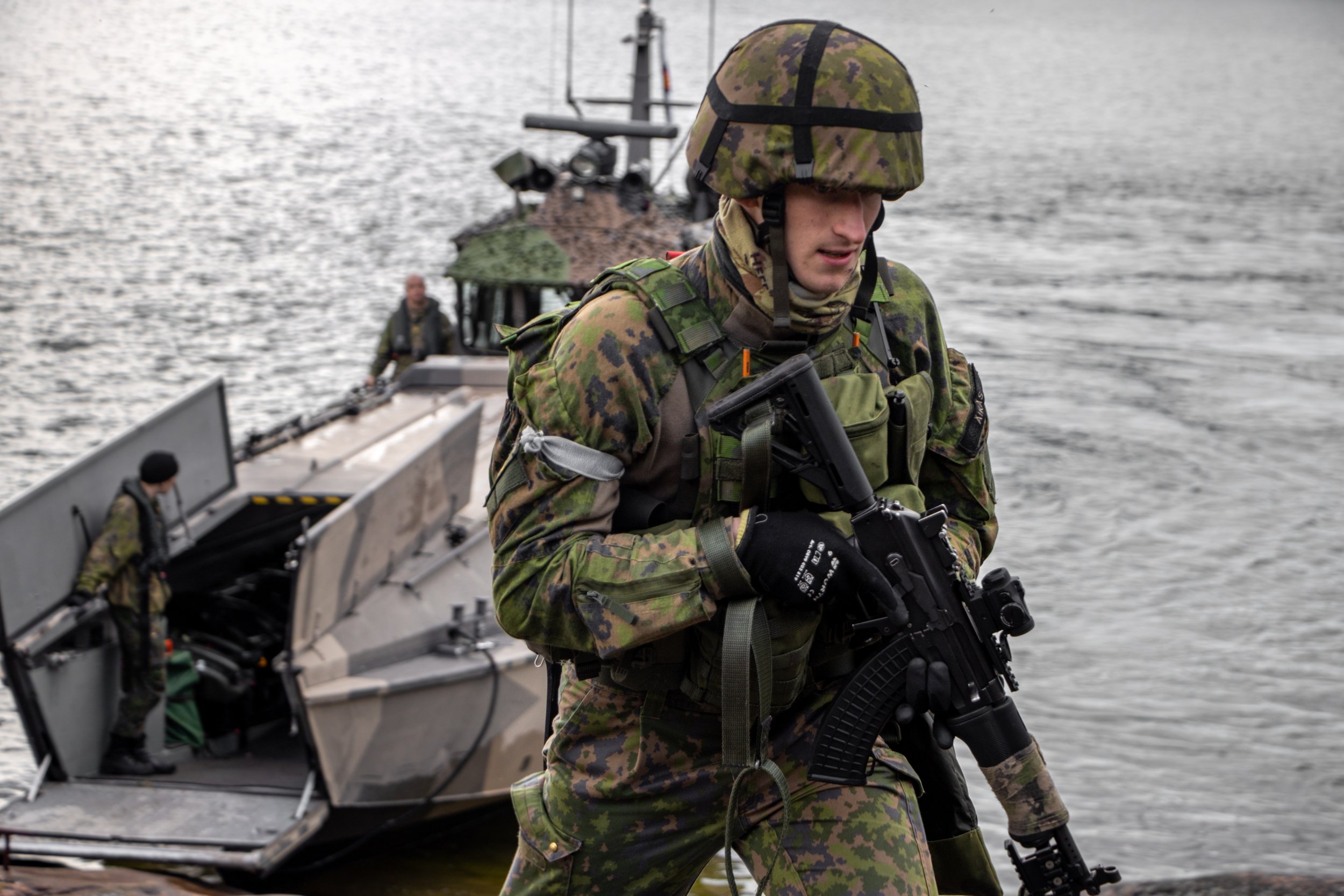 A soldier in camouflage uniform and helmet, holding a rifle, walking near a body of water with a military boat in the background.