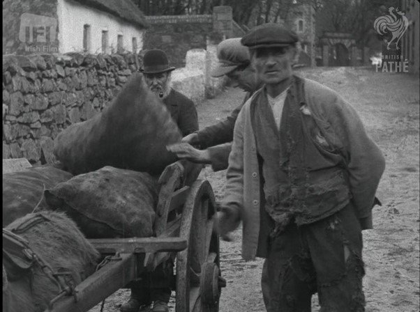 Black and white photo of men loading sacks onto a cart in a village street, with stone walls and thatched cottages in the background.