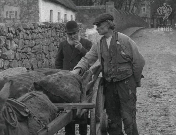 Two men in vintage clothing stand by a horse-drawn cart loaded with burlap sacks on a cobblestone street, with stone walls and rustic houses in the background, captured in a black and white photo.