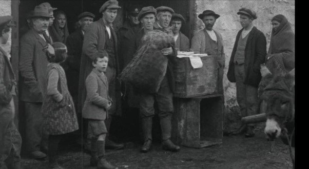 Black and white photo of people in old-fashioned clothing, standing around a box and holding a large bag, possibly engaged in a transaction or distribution, with a donkey partially visible on the right.
