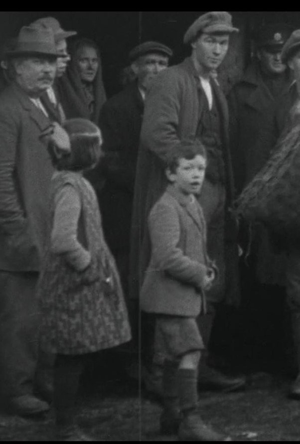 Vintage black and white photo of people, including children and adults, dressed in early 20th-century clothing, with hats and coats, standing outdoors in a group.