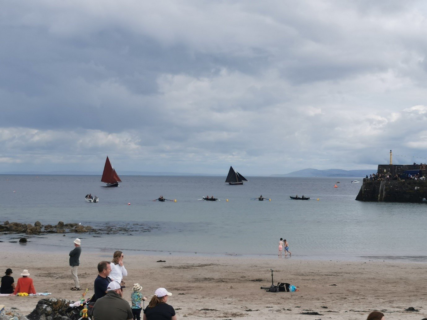 People sitting and standing on a beach with small boats and kayaks in the water, a stone pier visible to the right, and cloudy skies overhead.