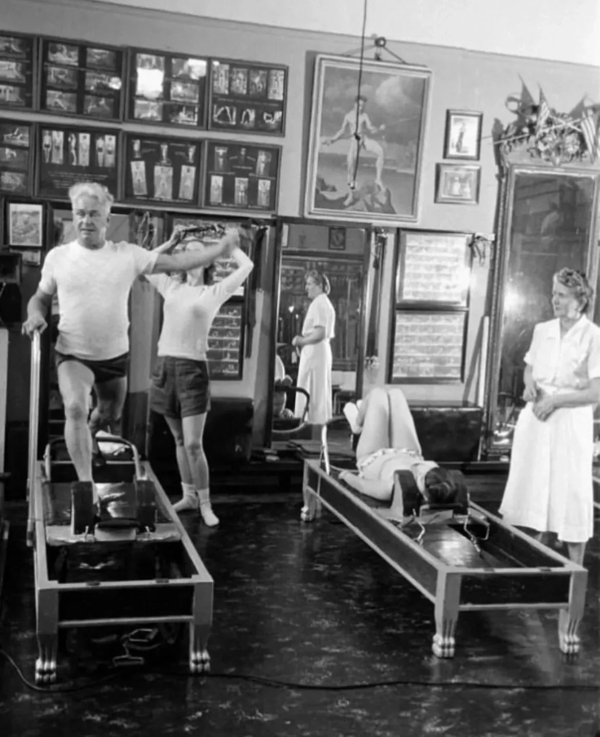 Black-and-white photo of fitness pioneer Joseph Pilates instructing students on reformer machines in a vintage Pilates studio, surrounded by mirrors, exercise charts, and framed artwork.