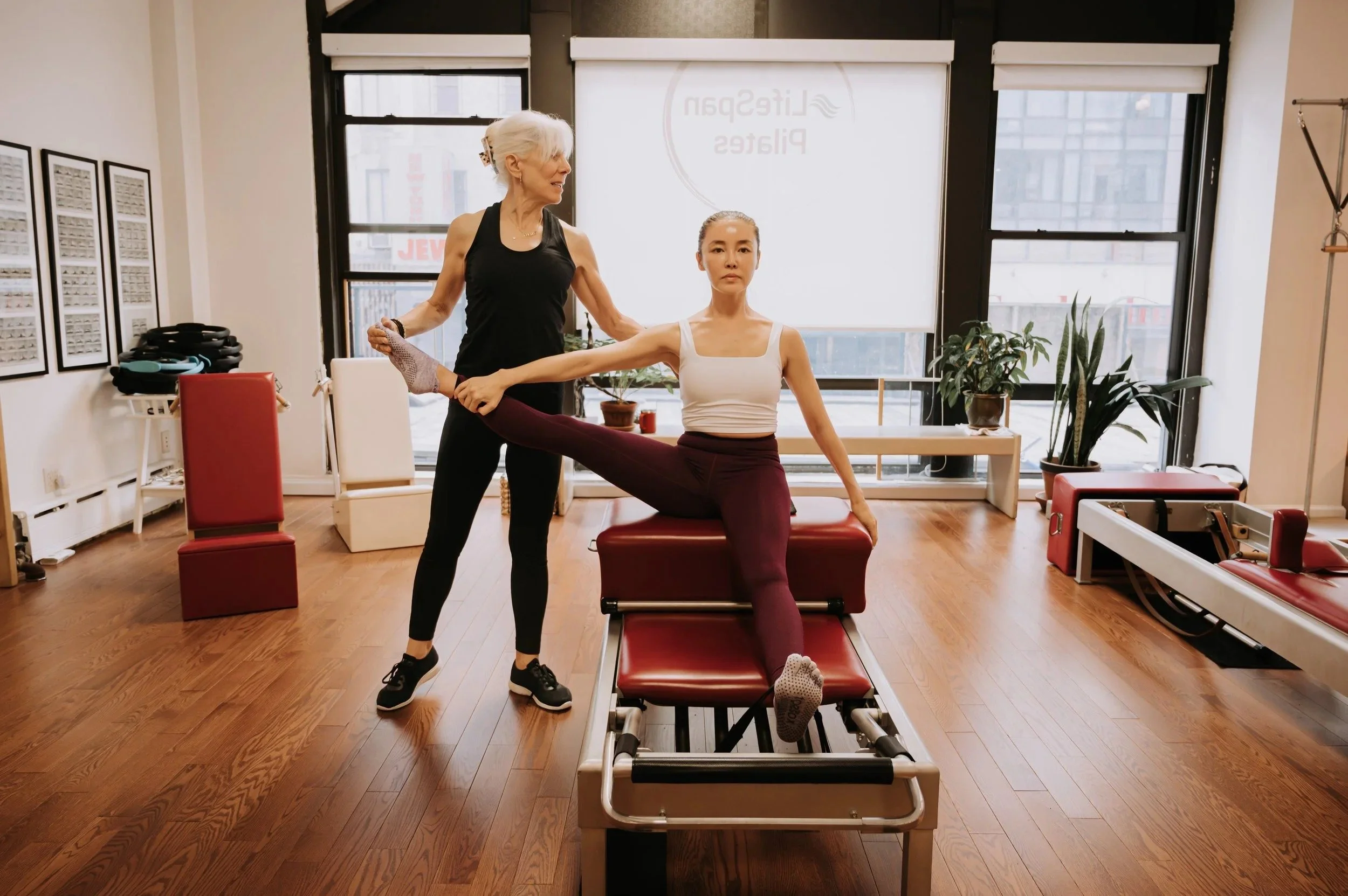 A woman does a exercise on a red Gratz Pilates reformer while an older instructor helps align and support her extended leg in a bright studio.