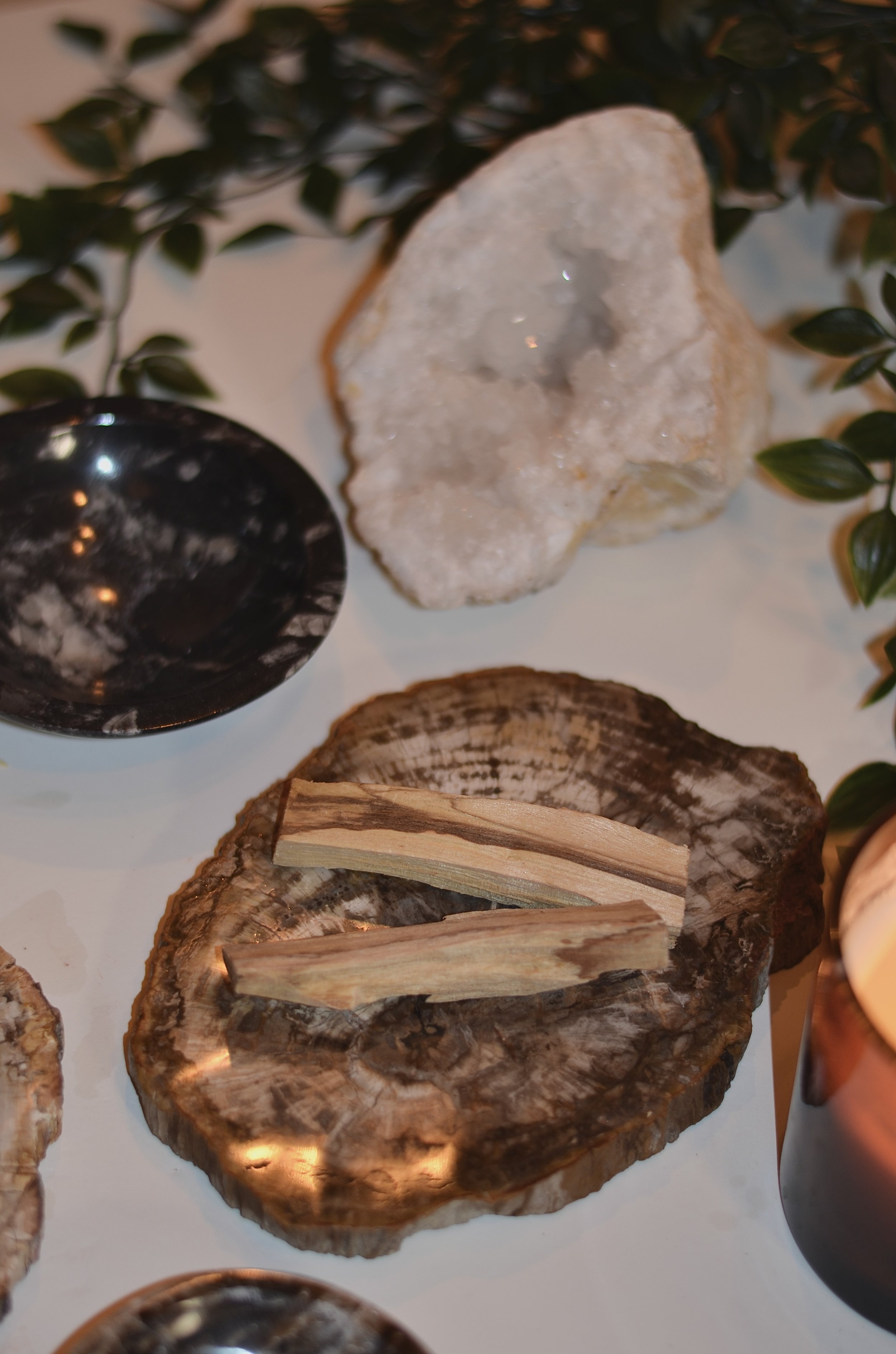 Weiße Kristallgeode und Palo Santo Holzstückchen auf Holzscheibe, schwarze Schale und grüne Blätter im Hintergrund.
