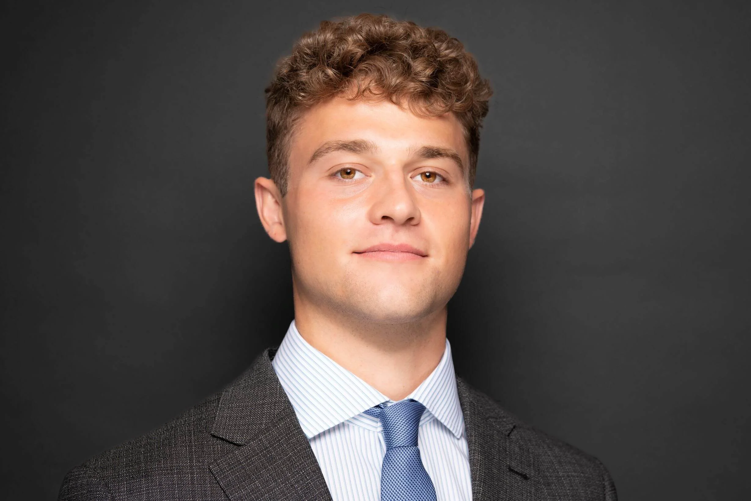Young man in a business suit with a blue tie, standing against a dark background.