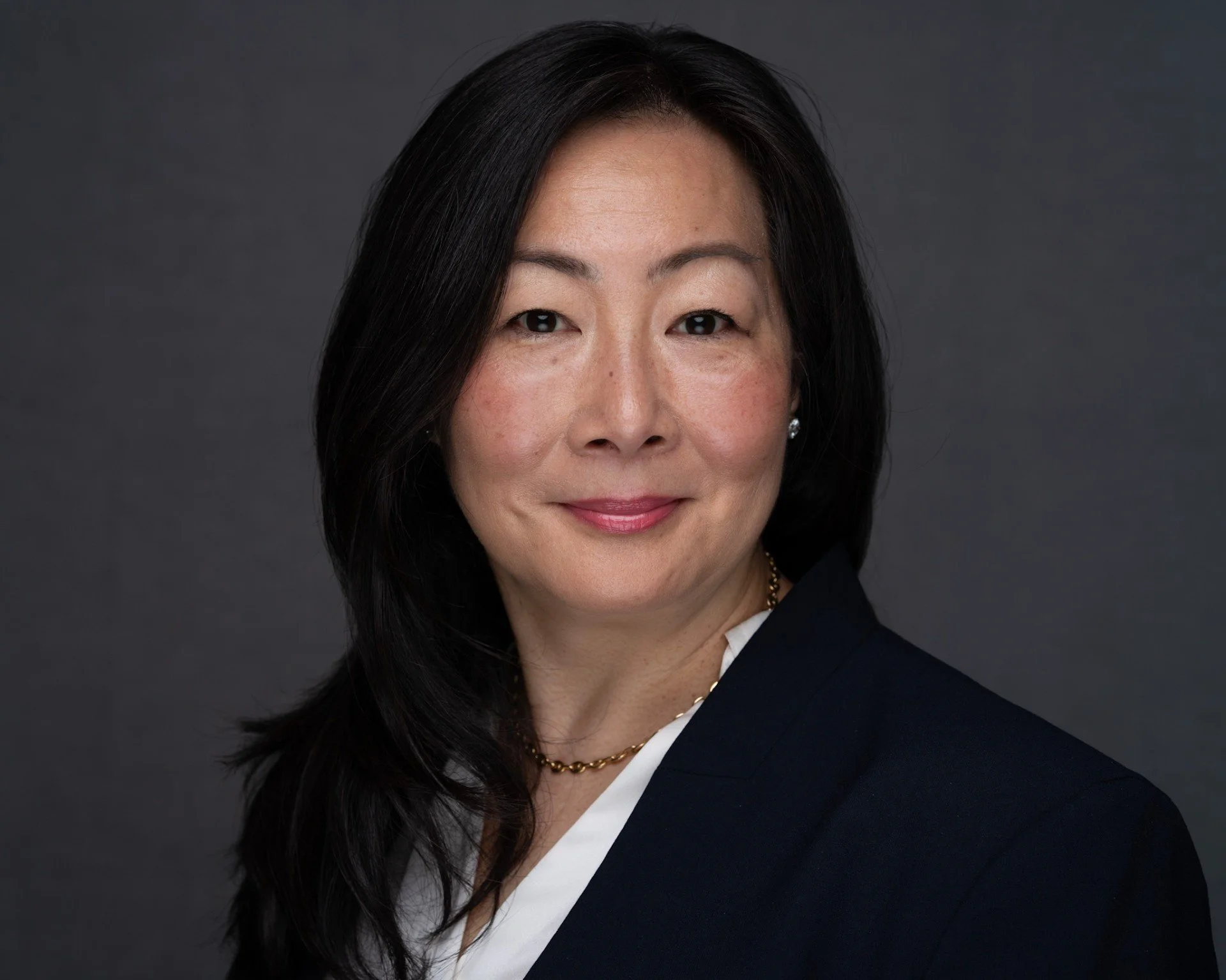 A professional headshot of an Asian woman with shoulder-length black hair, wearing a navy blazer and white shirt, against a dark gray background.