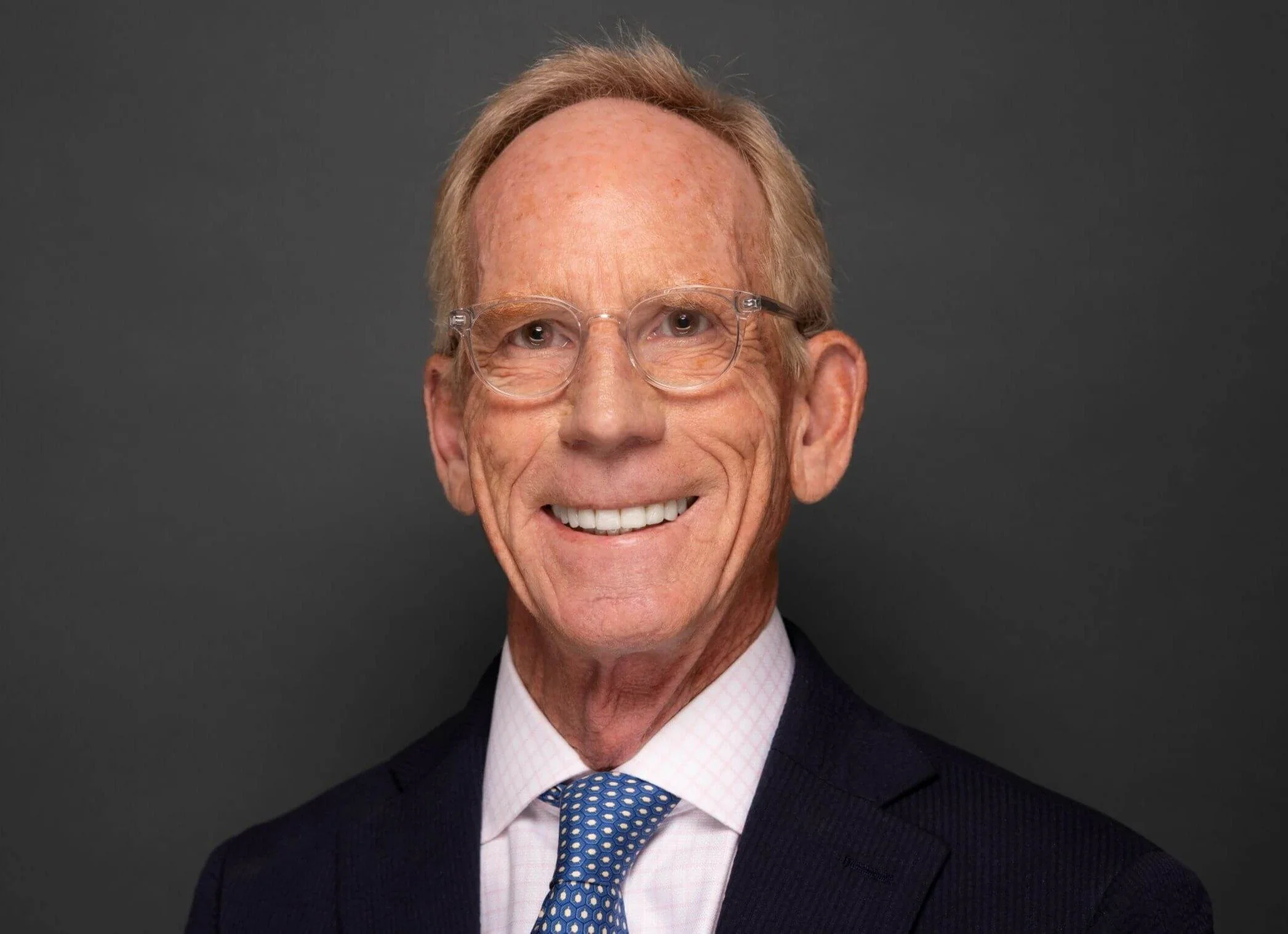 Portrait of an older man with glasses, short blond hair, wearing a navy suit, pink checkered shirt, and blue patterned tie, smiling against a dark gray background.
