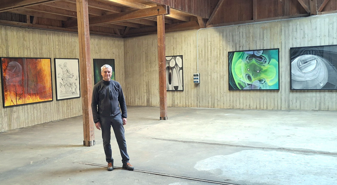 Der Künstler Martin Schmidt steht in einer hölzernen Halle (Kugelhalle des Künstlerhauses Ganslberg). An den Wänden sind mehrere seiner Malereien ausgestellt.