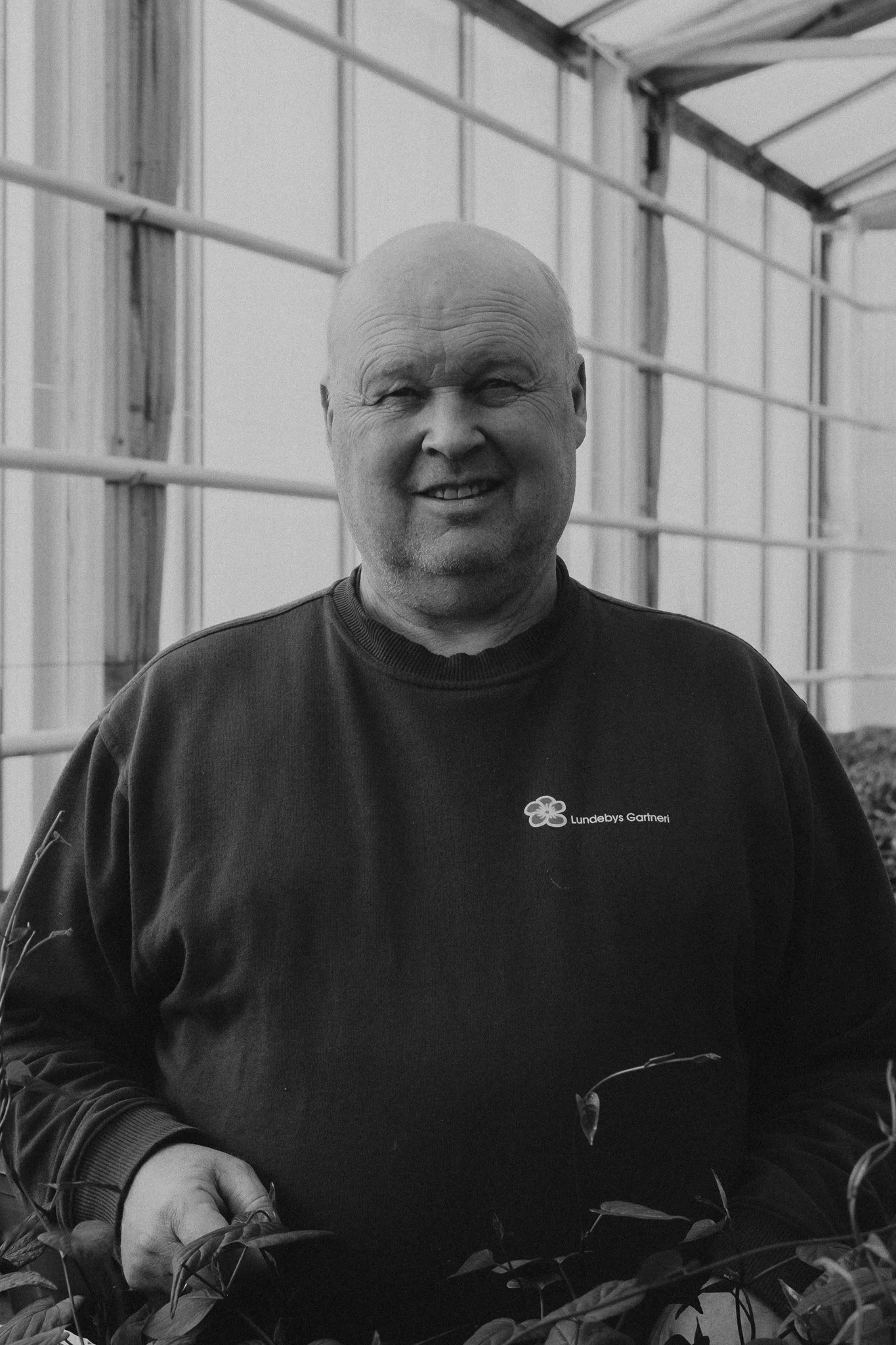 En smilende mann med skjegg i en drakt, inne i et drivhus med grønne planter rundt ham.