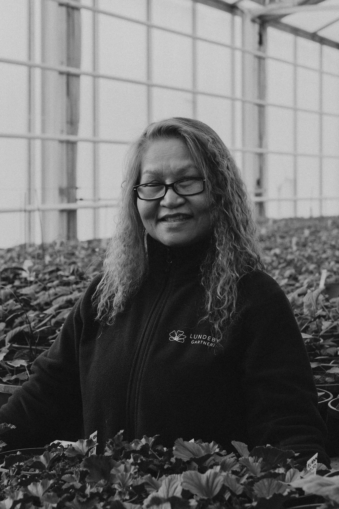 En kvinne med briller smiler inn i kameralinsen, hun står i et drivhus med mange planter rundt seg.