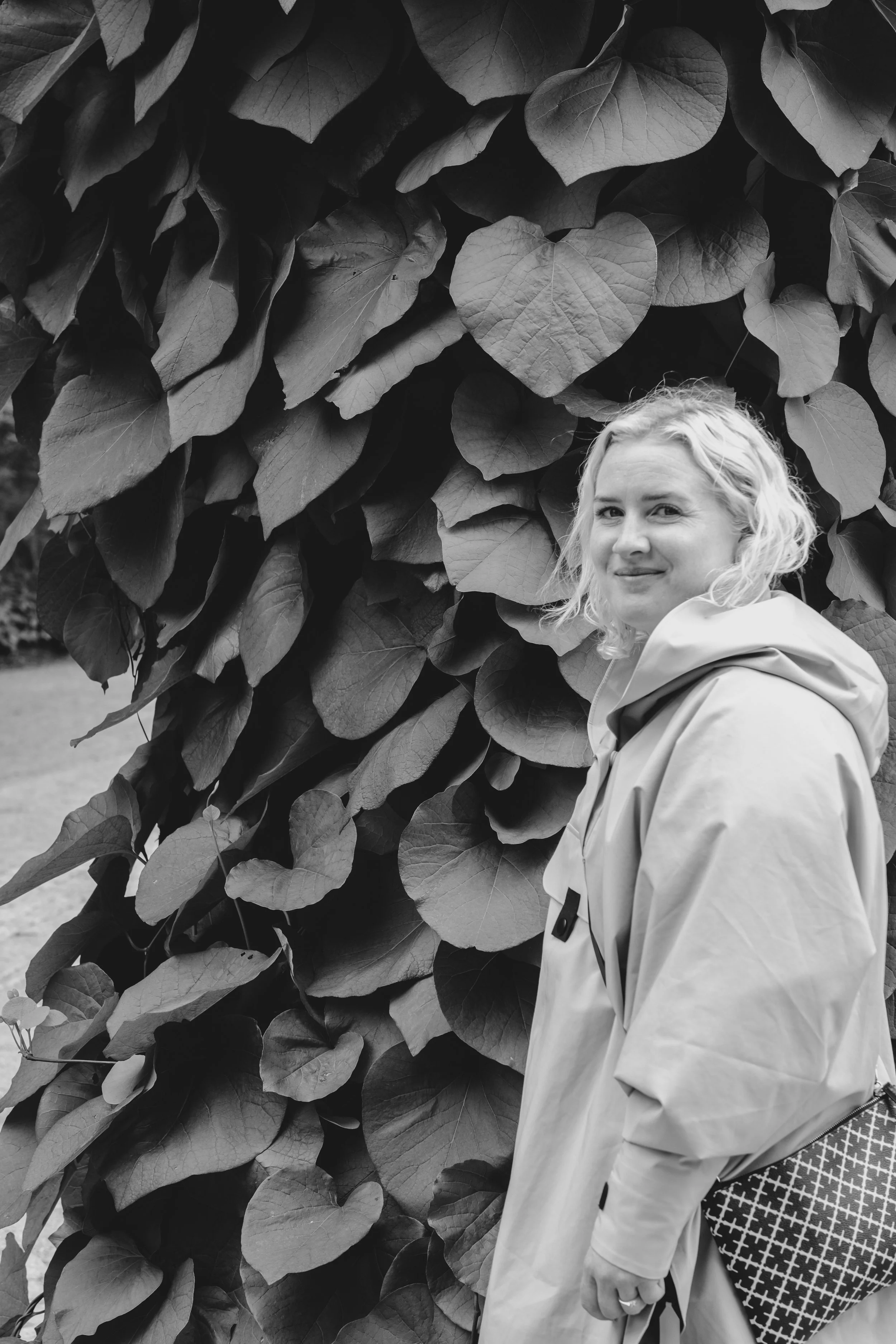 Et svart-hvitt bilde av en kvinne med lyst, krøllete hår som står foran en vegg av store blader.