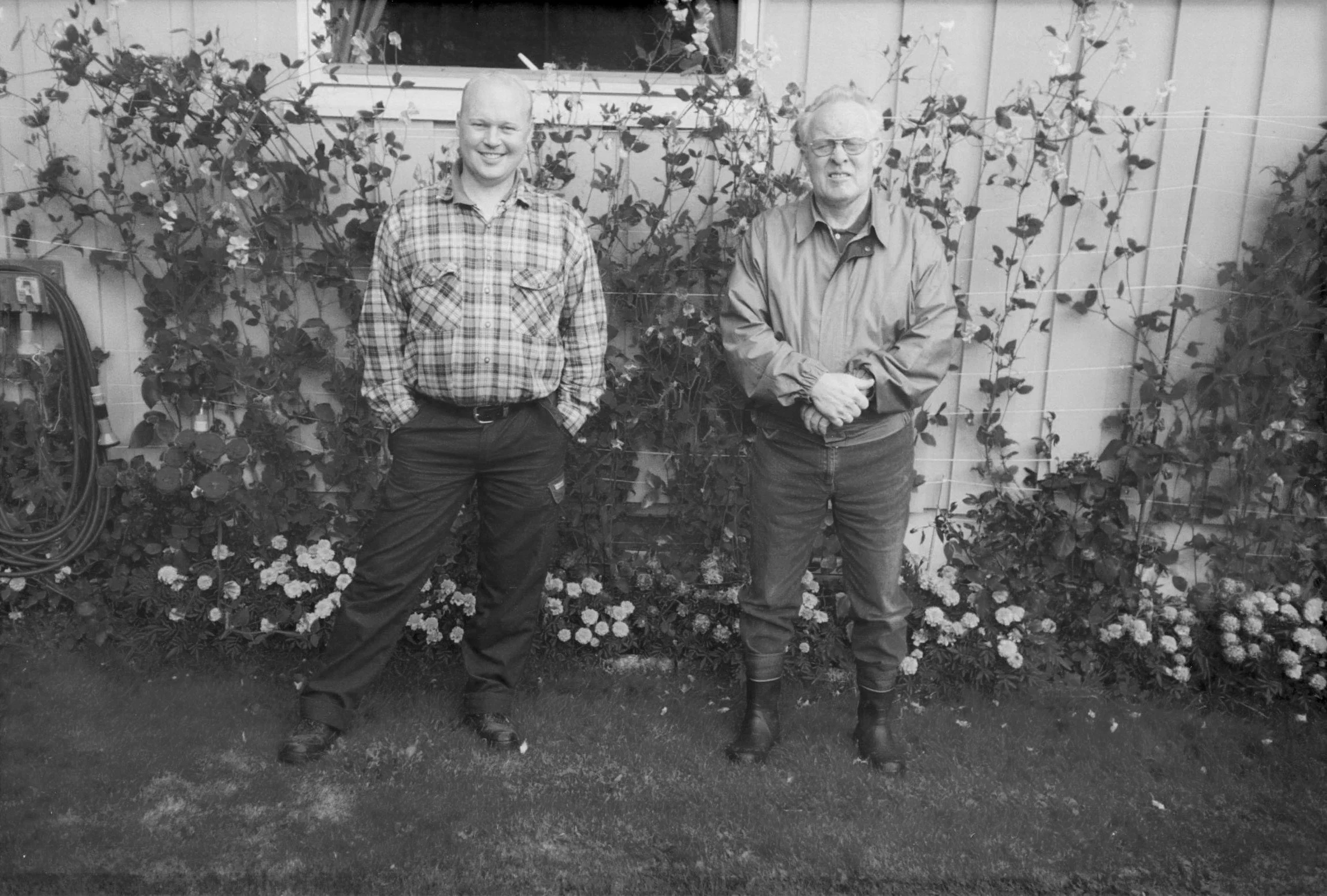 To menn står i en hage foran en hekk med blomster. Den ene mannen har på seg en sjakett og har hendene i lommene, mens den andre mannen har på seg en jakke, og har hendene foldet foran seg. Det er en vegg og et vindu bak dem.