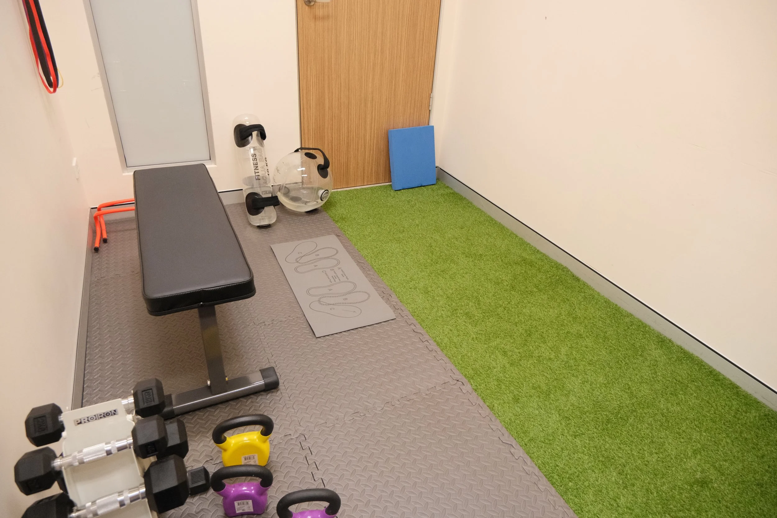 Home gym corner with a black workout bench, colorful kettlebells, a water bottle, a kettlebell, a blue foam pad, a glass medicine ball, an exercise mat with the word 'GET UP' on it, and a patch of artificial green turf on the floor.