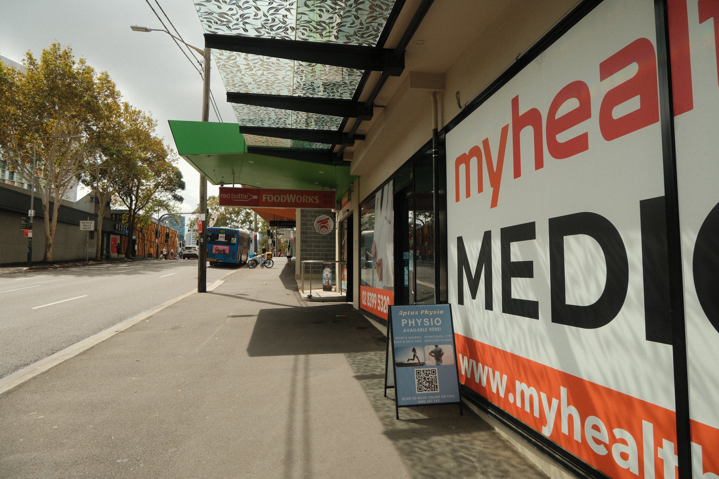 Street view with a pharmacy storefront on the right, featuring a large sign with 'myhealth' branding, and a sidewalk in front. There is a bus and bicycle parked on the street, trees lining the sidewalk, and advertisements including a sign for physiot