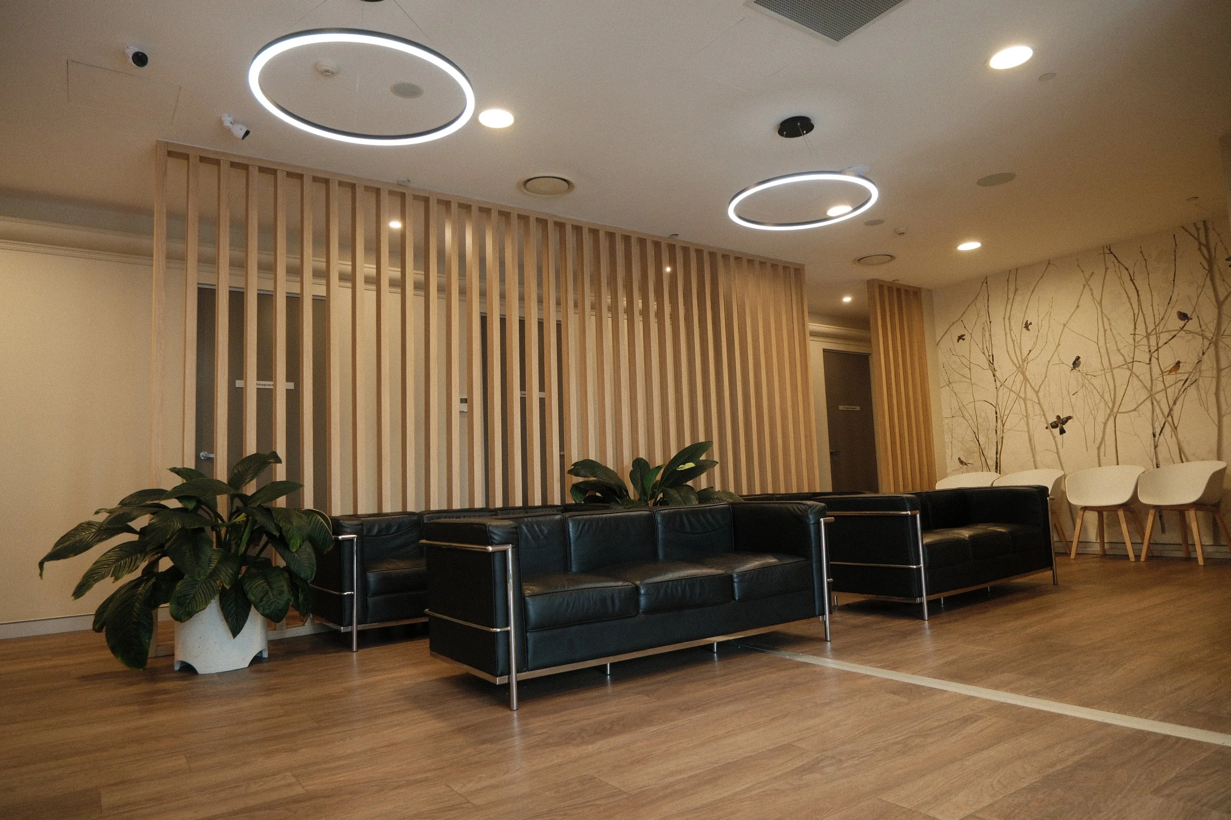 Modern waiting room with black leather sofas, white chairs, potted plants, circular ceiling lights, and artwork of trees and butterflies on the wall.